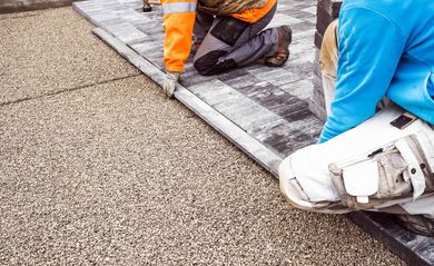 Workers installing paving stones next to a gravel path, with a white machine and orange work pants visible Workers installing paving stones next to a gravel path, with a white machine and orange work pants visible