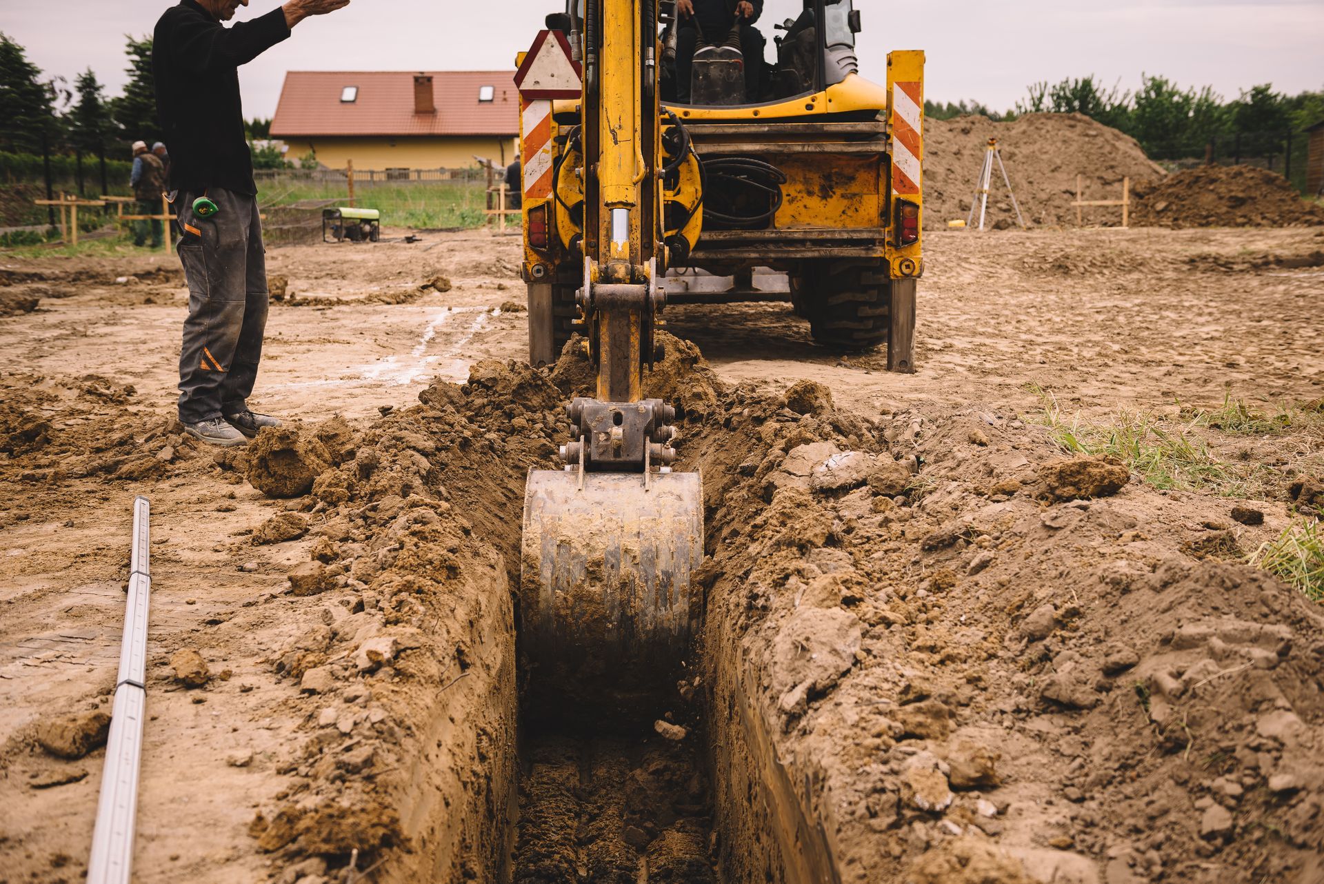 Backhoe digging a trench at a muddy construction site with a worker nearby