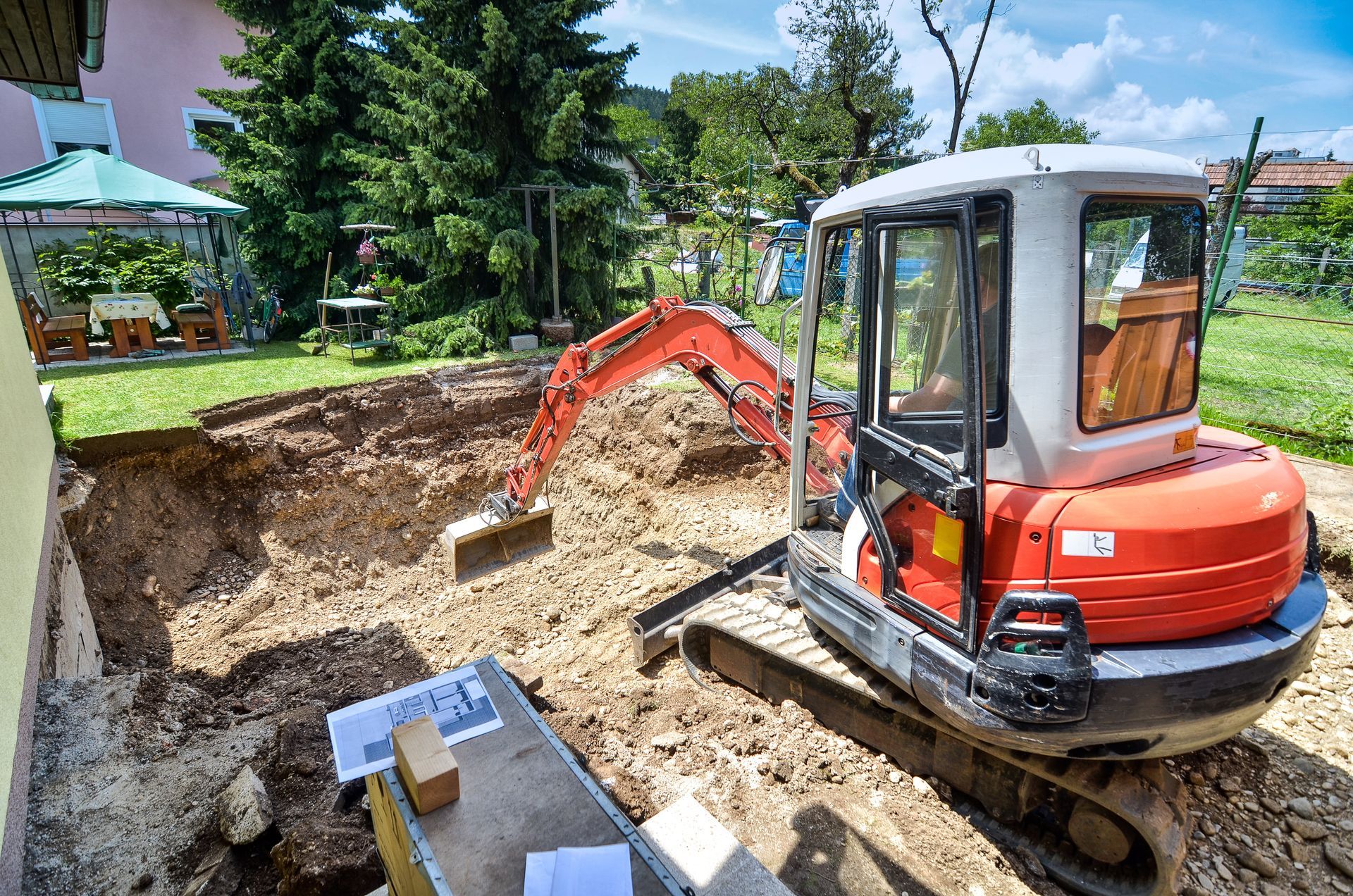 Red mini excavator digging a backyard trench beside a house and trees