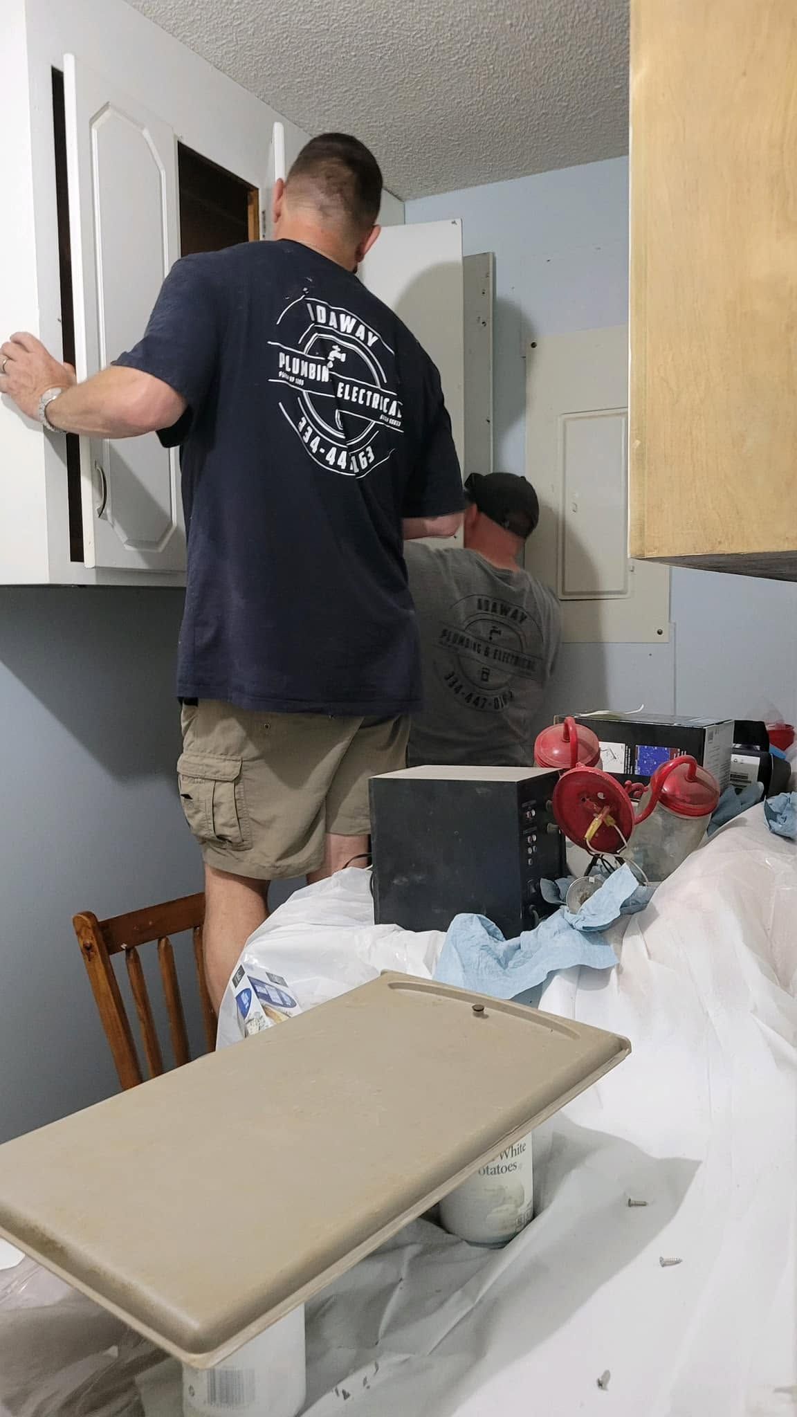 Two people working in a small kitchen, one standing by the door and one kneeling near the cabinets.