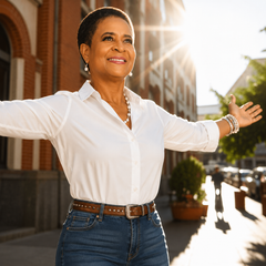 Smiling person in white shirt with arms outstretched on a sunny city sidewalk.