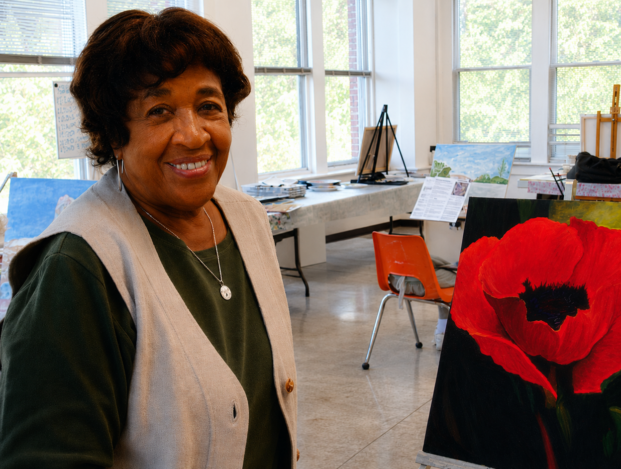 Smiling woman in a bright room beside a large red flower artwork, with tables and chairs behind her