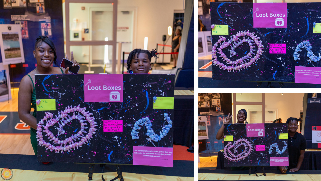 Two smiling youth stand behind poster using lollypops to visualize data about loot boxes