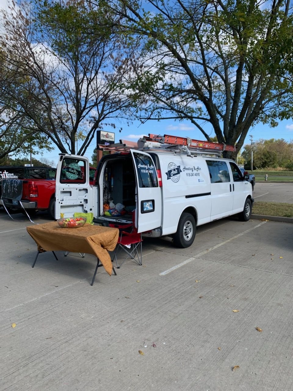 White van with rear doors open, parked under trees beside a folding table and bench in a lot.