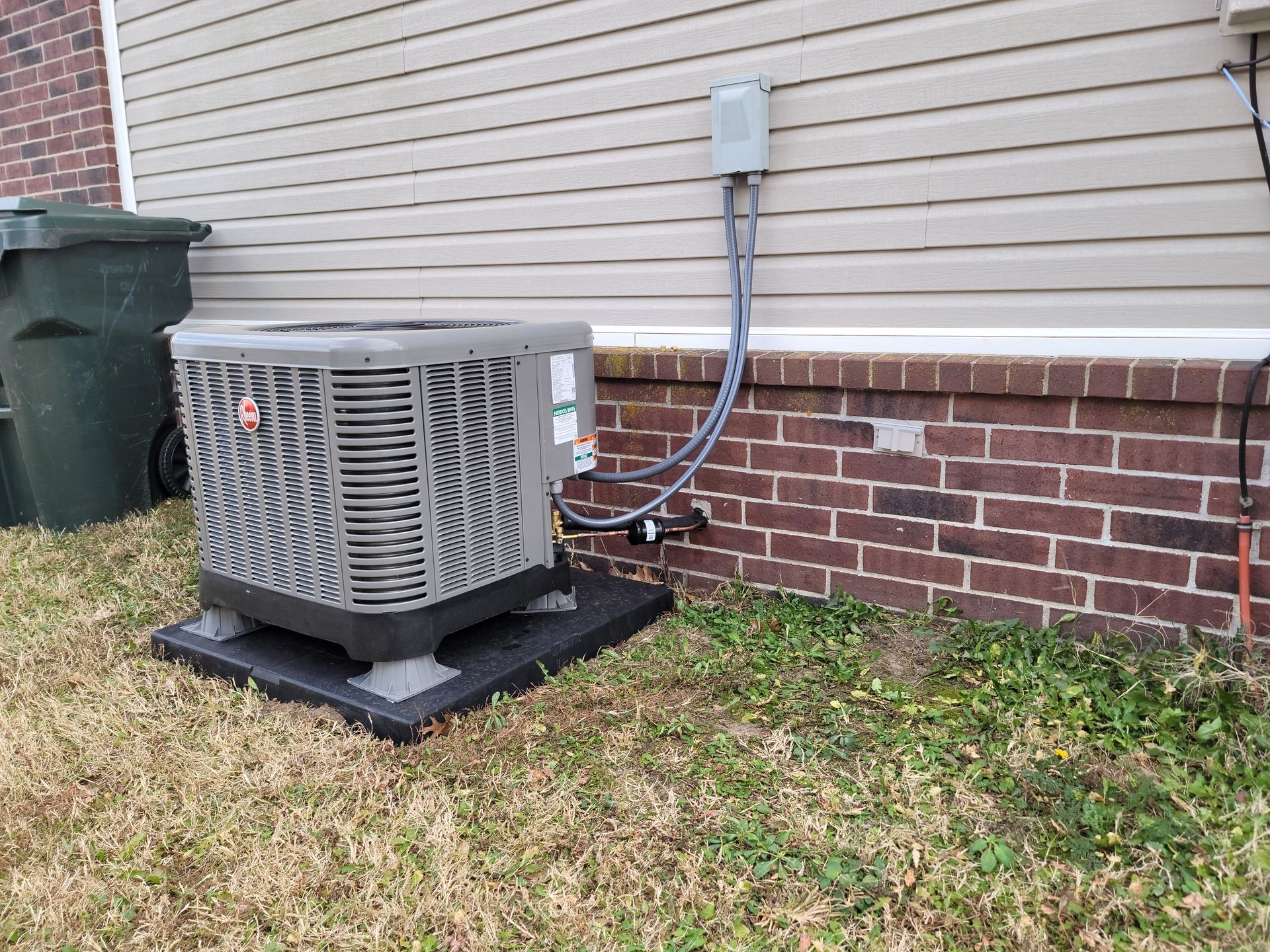 Outdoor air conditioner unit beside a beige house with brick foundation and utility conduit