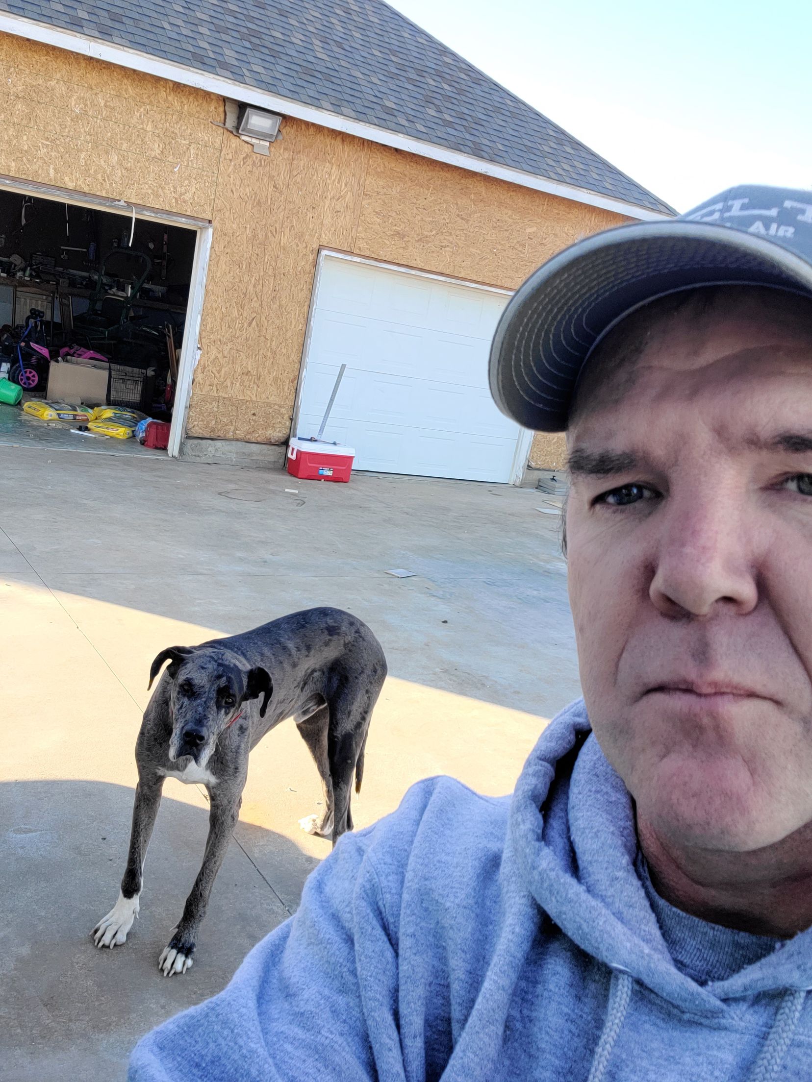Man taking selfie beside a spotted dog in a driveway, with an open garage and brick house behind.