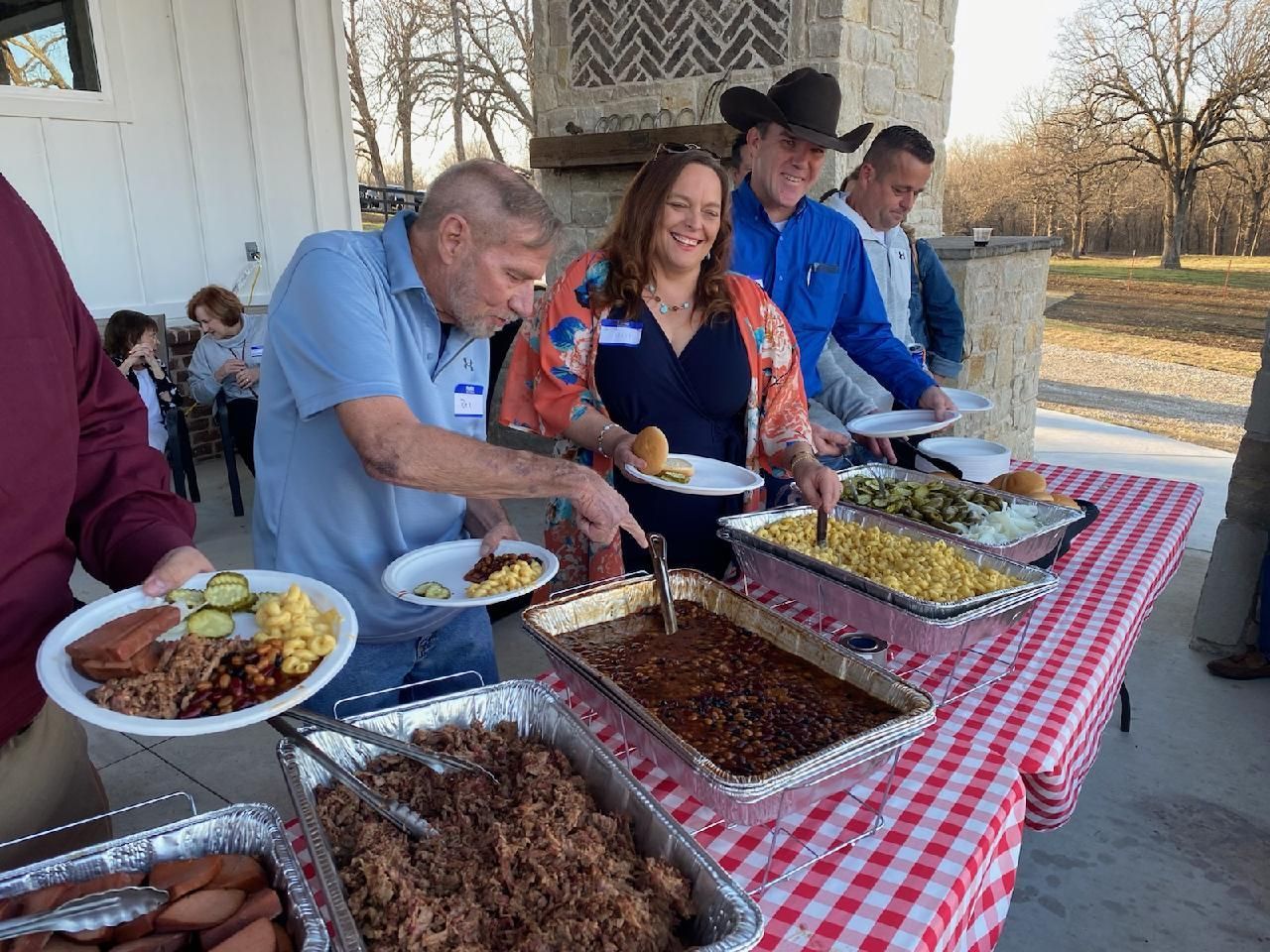 People serving food at a buffet table under a porch, with trays of dishes and cups.