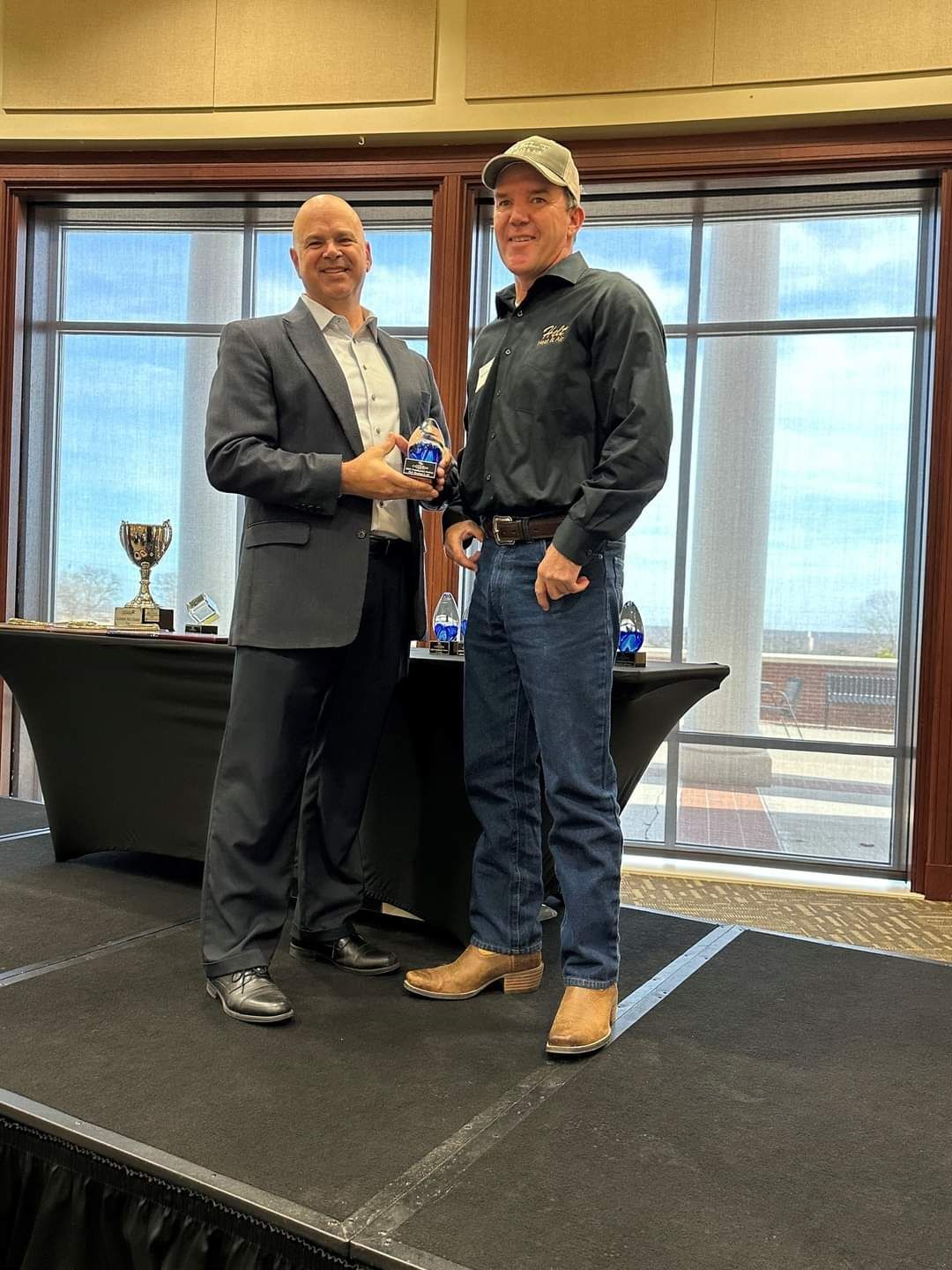 Two men standing in a conference room, one holding a small trophy, with city windows behind them.