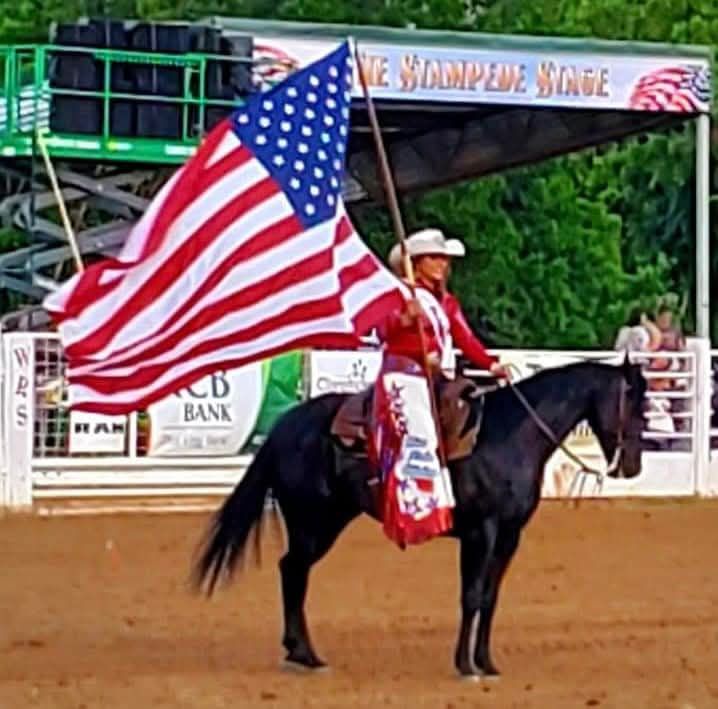 Cowgirl on a black horse carrying a large American flag in a rodeo arena