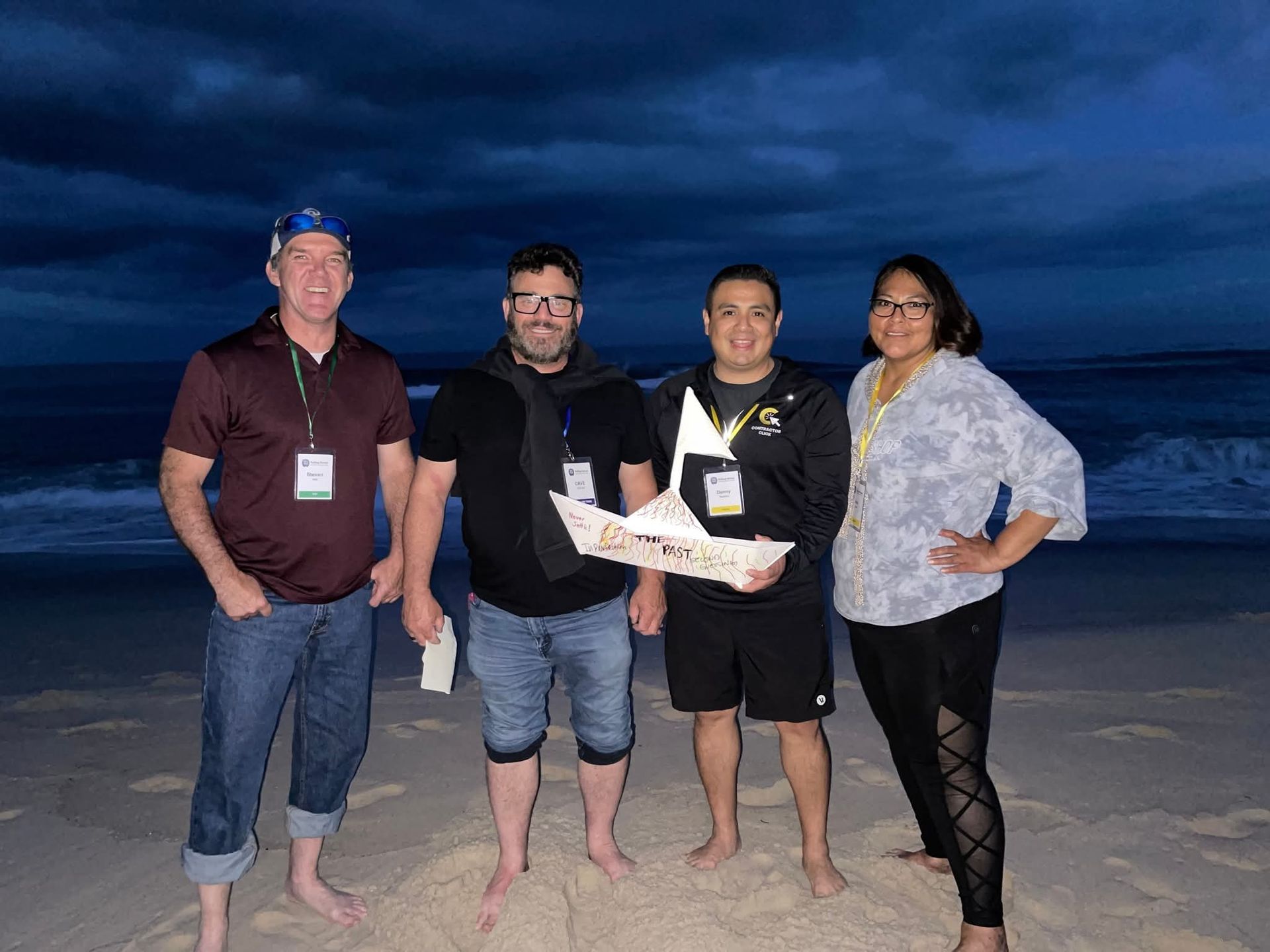 Four people standing barefoot on a beach at dusk, holding a sign and smiling at the camera.