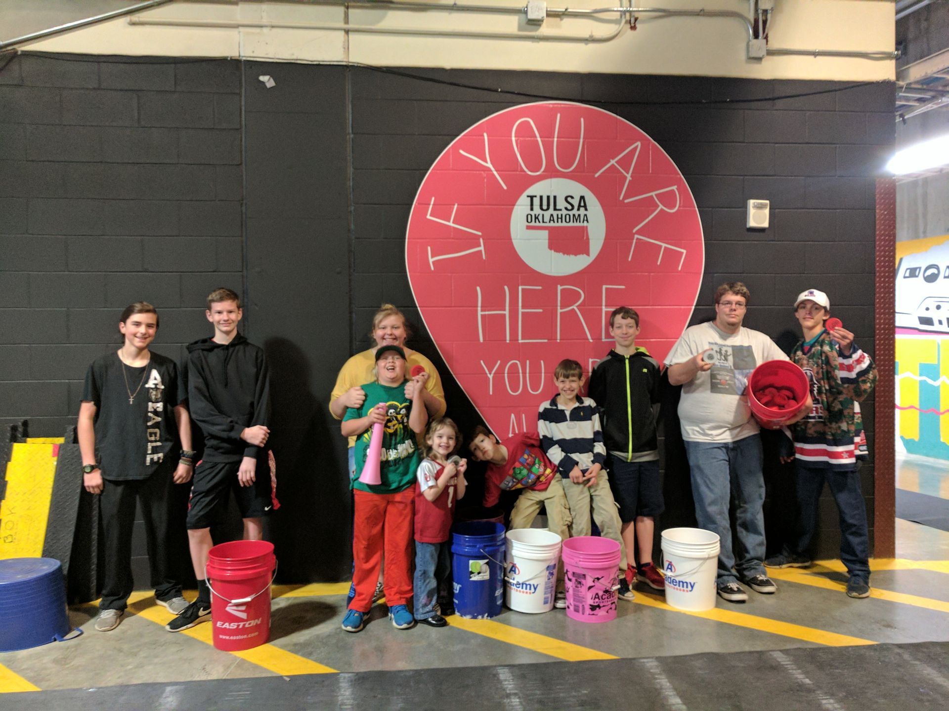 Group of people standing indoors in front of a large red “You are here” sign with colorful buckets on the floor.