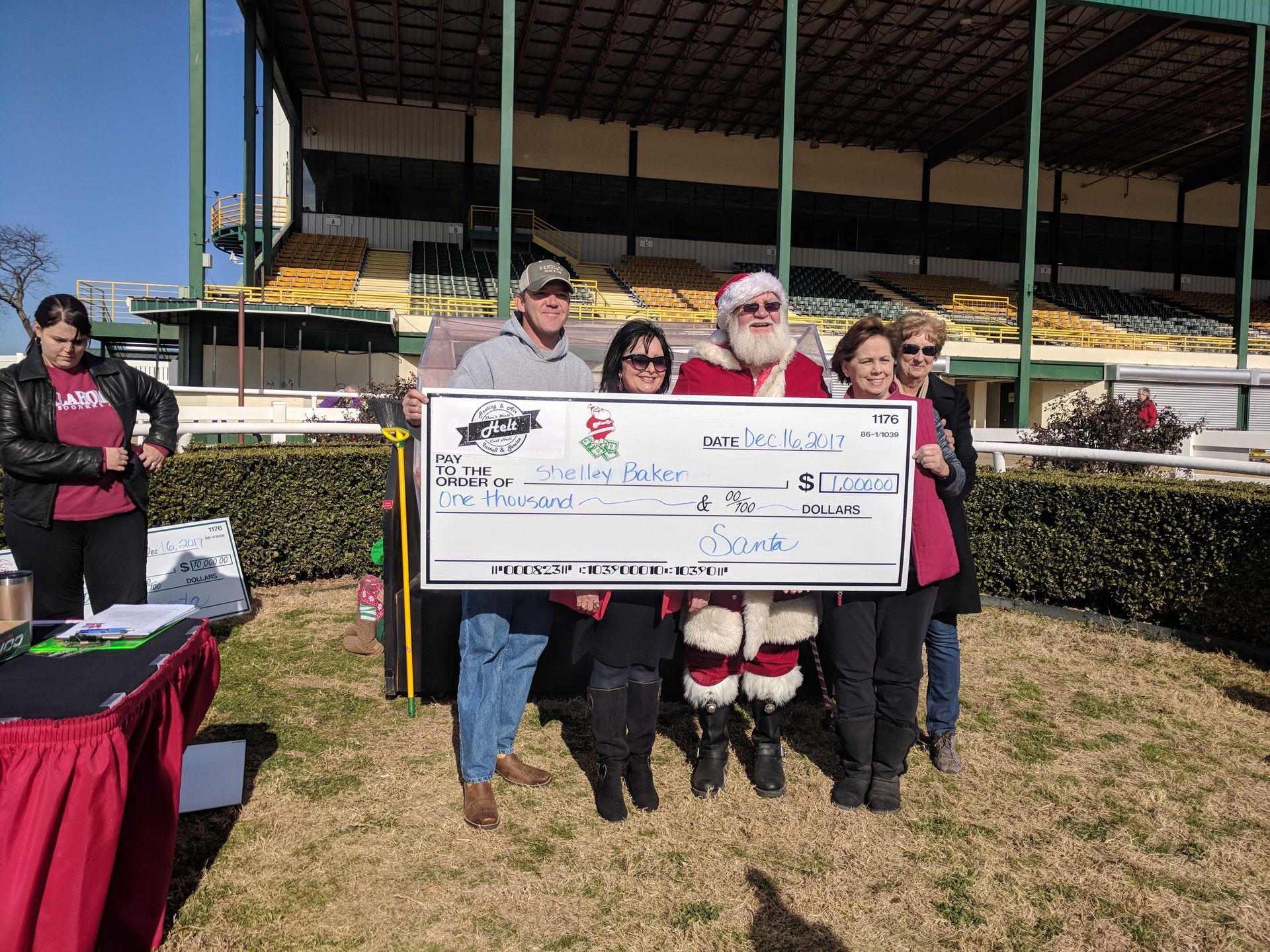 Four people holding an oversized check at an outdoor event, with a mascot in a Santa suit beside them.