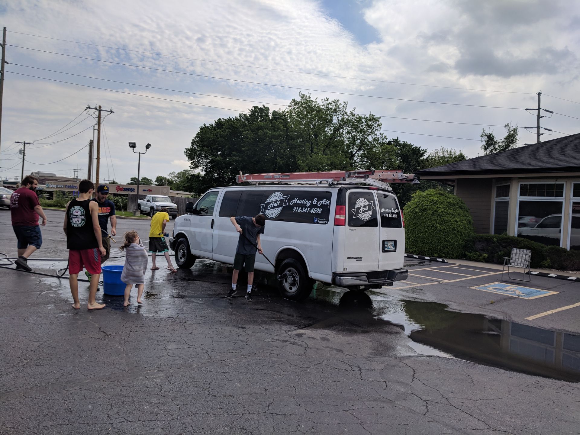People gathered around a white van on a wet street, with children playing near a puddle.