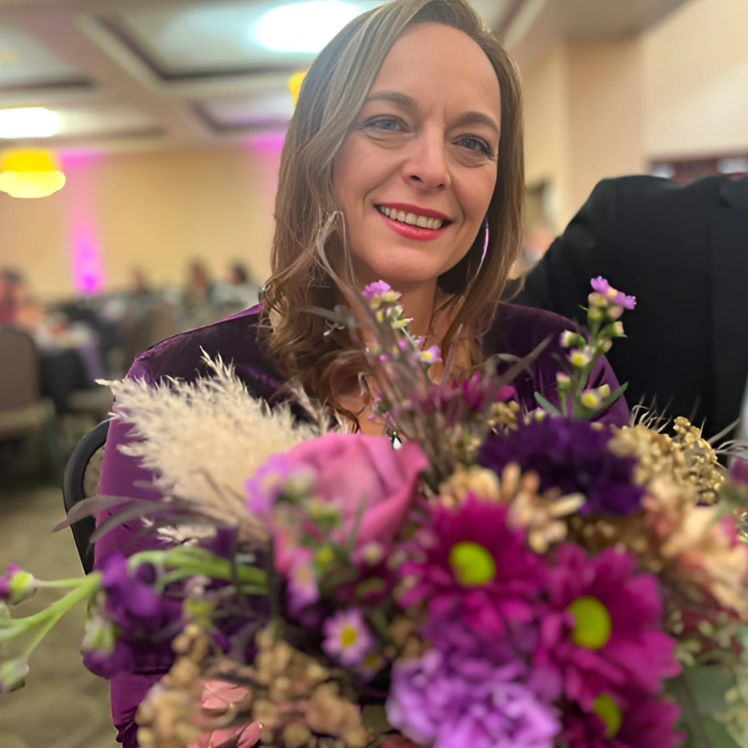 Woman smiling behind a colorful bouquet of purple and pink flowers at an indoor event