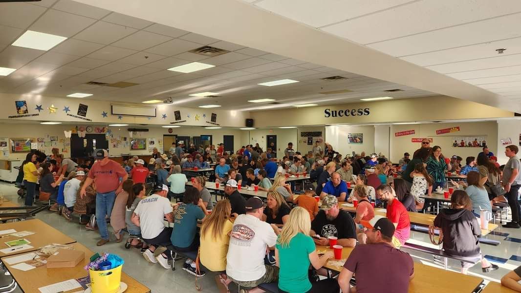 Crowded school cafeteria with many people seated at long tables eating and talking