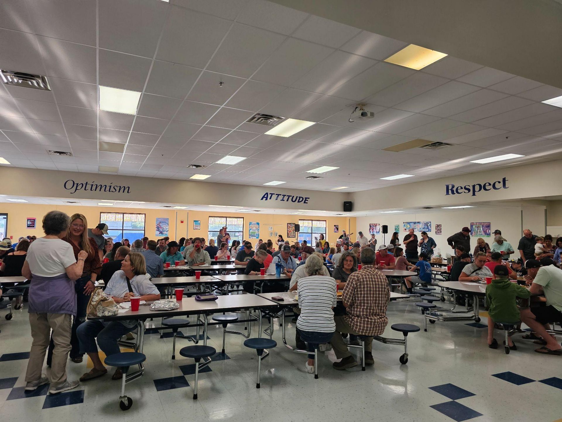 Busy school cafeteria with students seated at long tables under wall words “Question,” “Arrange,” and “Respect.”
