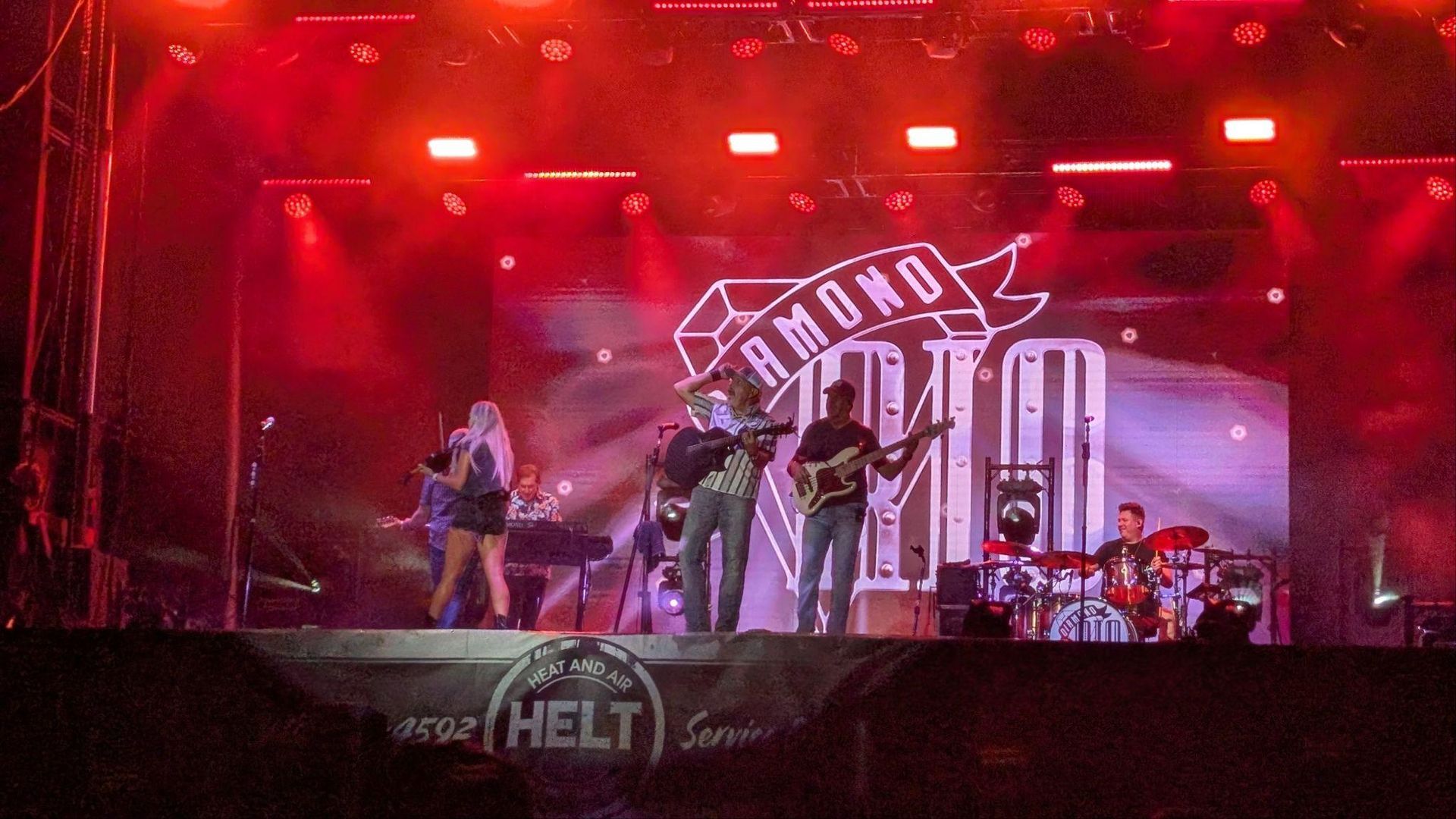 Rock band performing on a red-lit stage with drums and a large backdrop logo