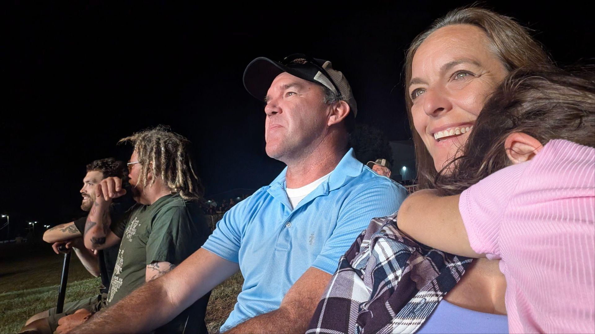 People seated outdoors at night, smiling and watching an event from bleachers.