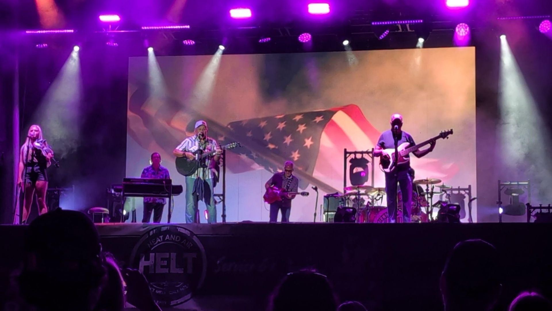 Band performing on stage under purple lights with a large American flag backdrop