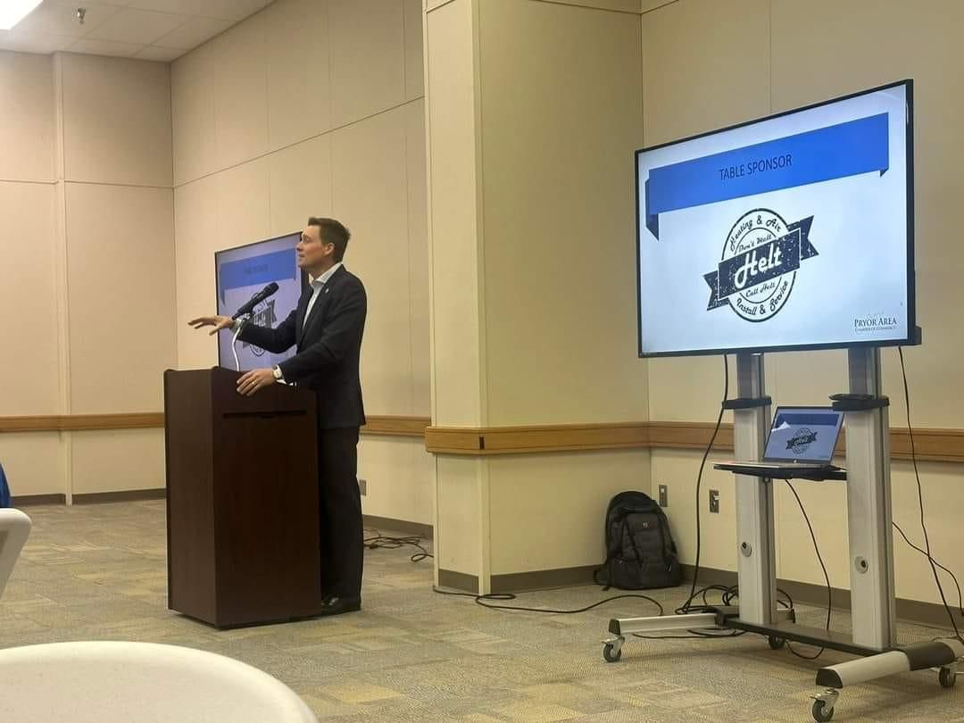 Man speaking at a podium in a beige conference room, with a presentation screen beside him.