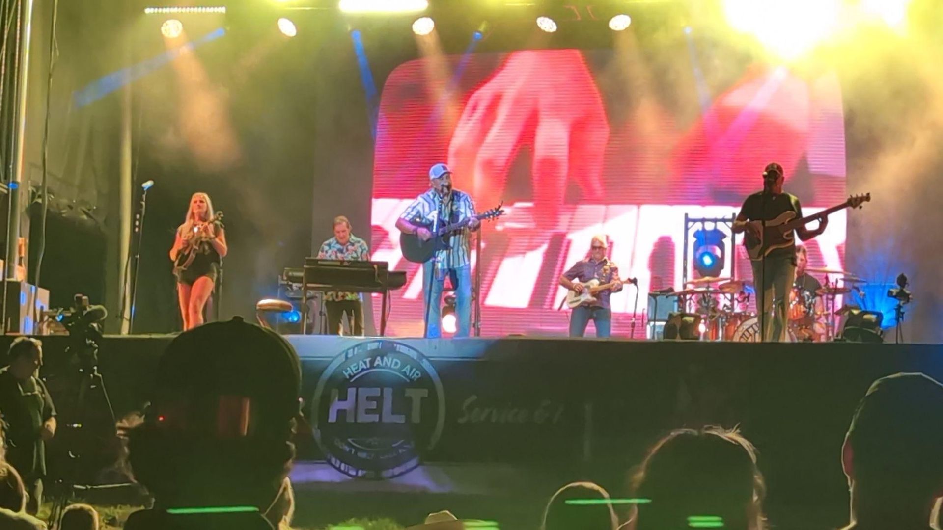 Live band performing onstage under colorful lights, with a large screen showing a close-up of hands playing keyboard.