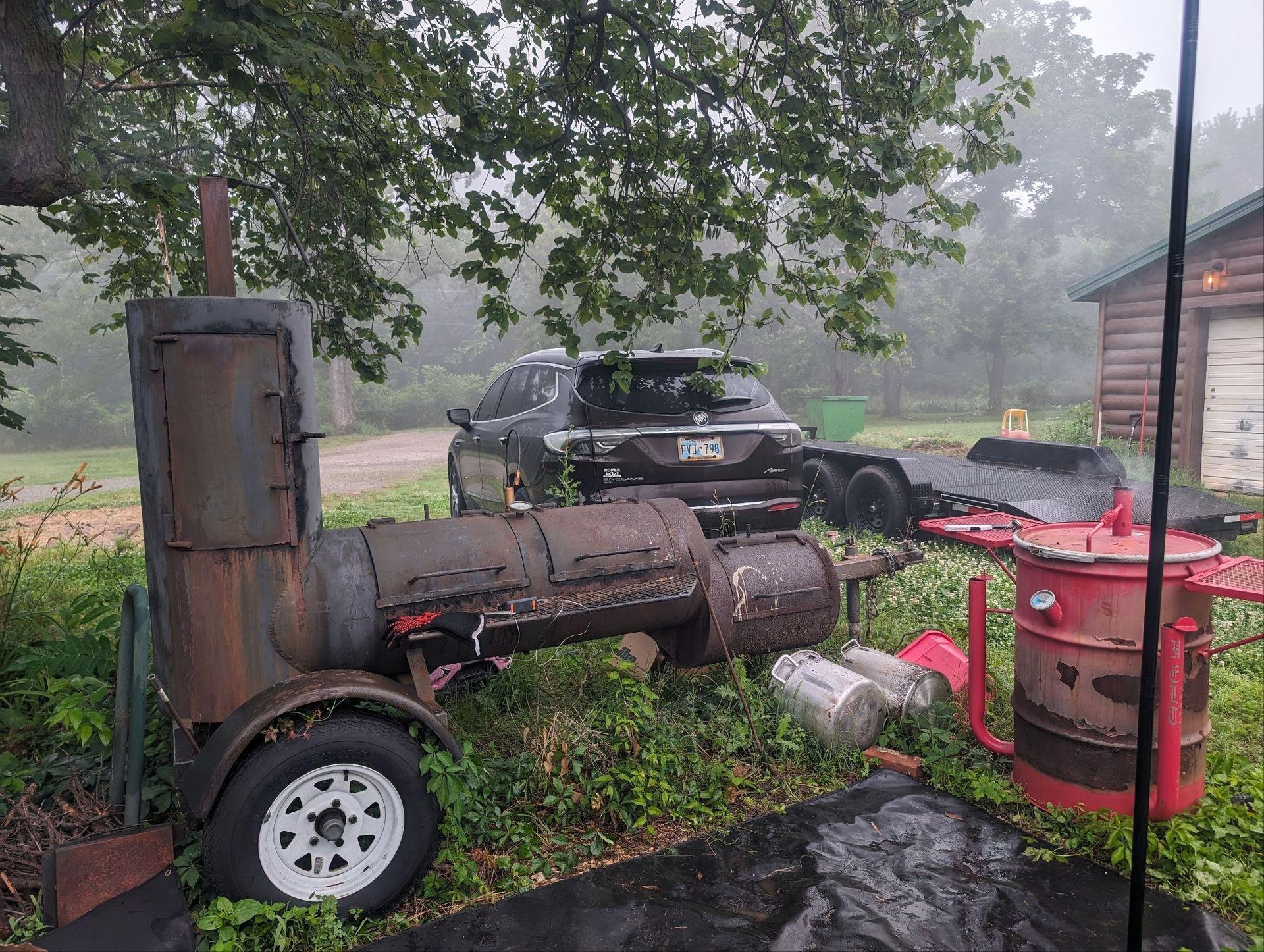 Rusty old trailer and grill beside a black SUV in a foggy yard with trees and a brick building