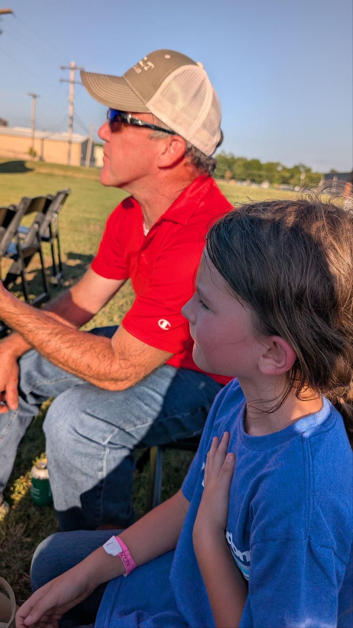 Two people sitting outdoors at sunset, one in a red shirt and cap, with chairs and open field in the background.