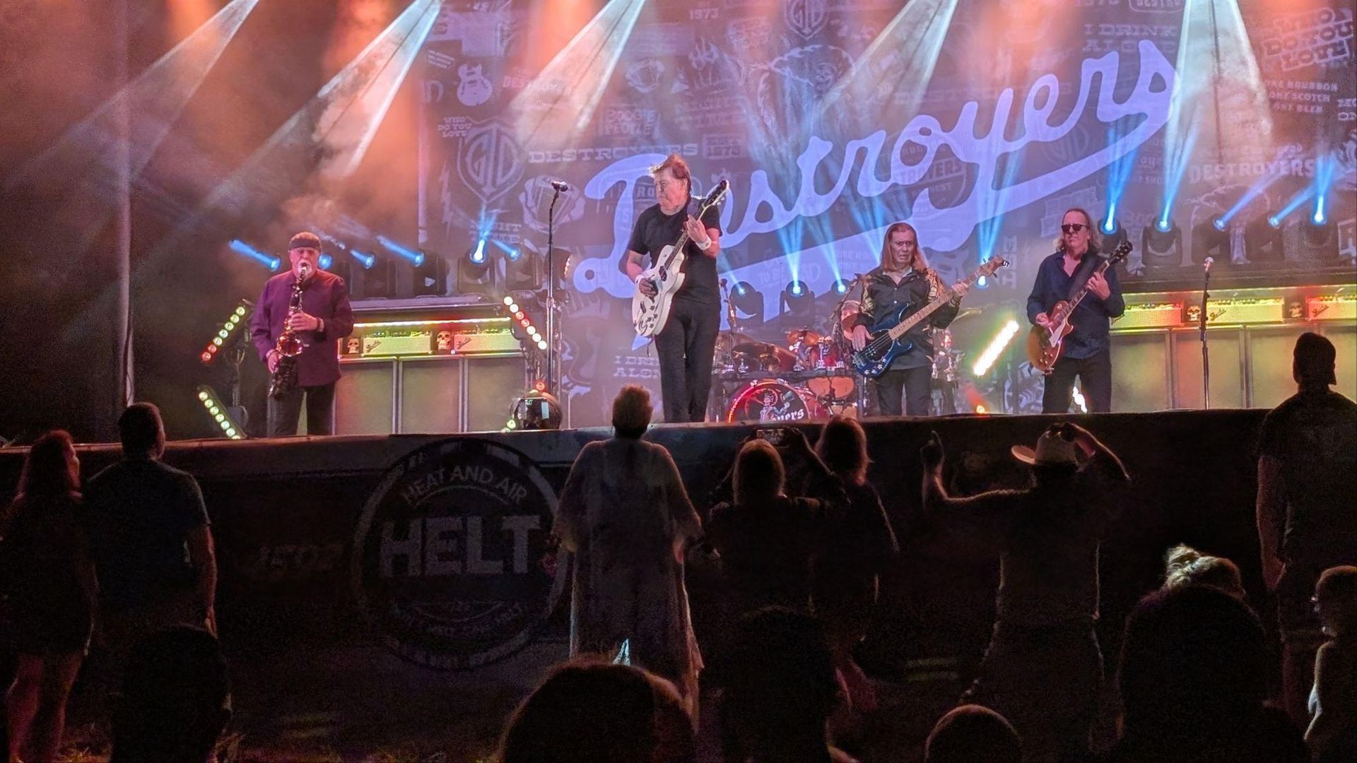 Rock band performing on a brightly lit stage with crowd silhouettes in the foreground