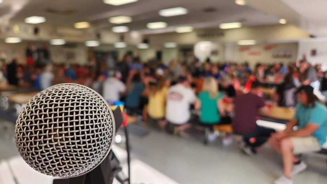 Microphone in foreground with a blurred crowd seated in a bright auditorium or classroom setting