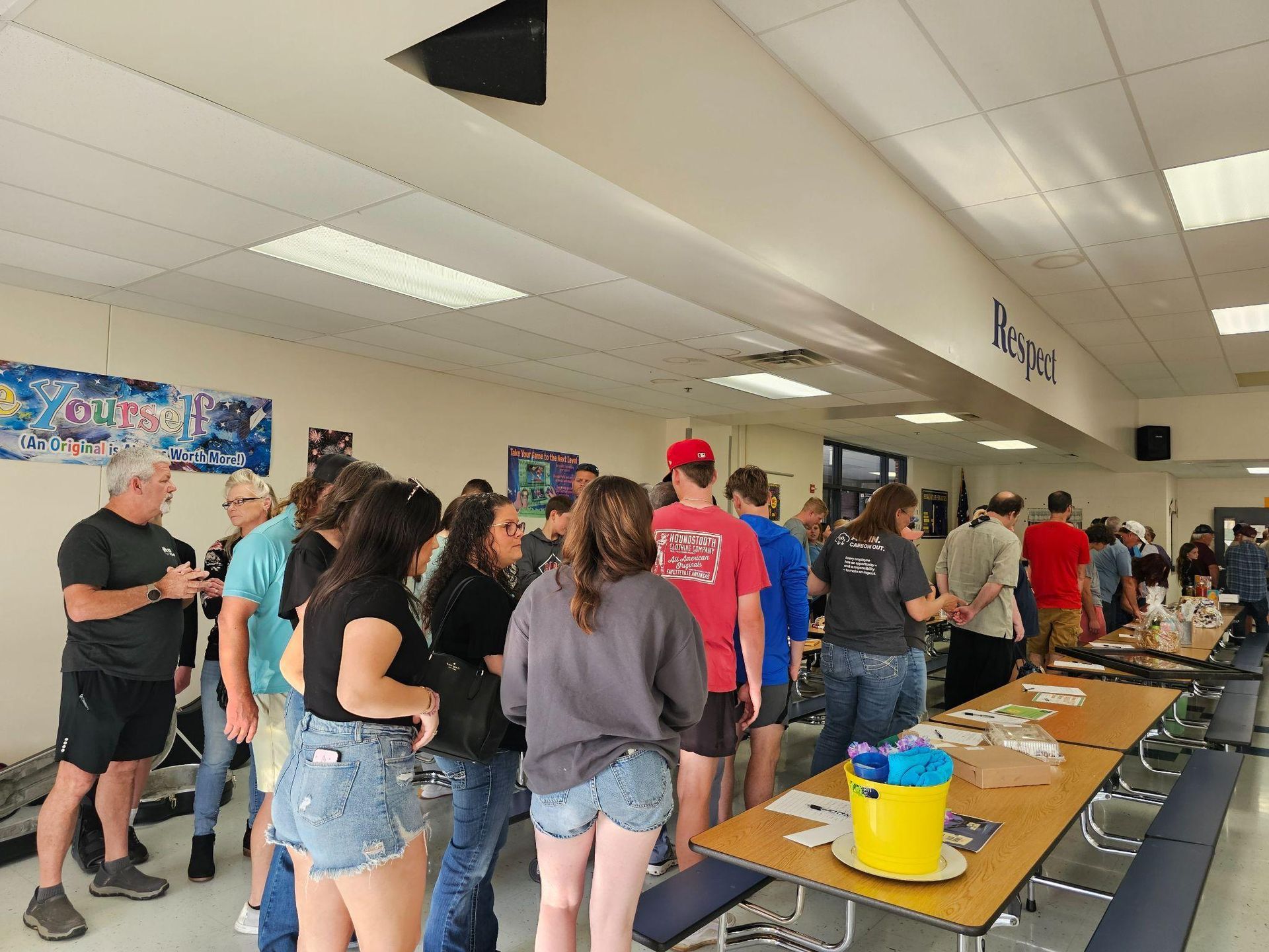 People standing in line at a cafeteria-style table in a school hallway, with trays, cups, and a yellow bucket.