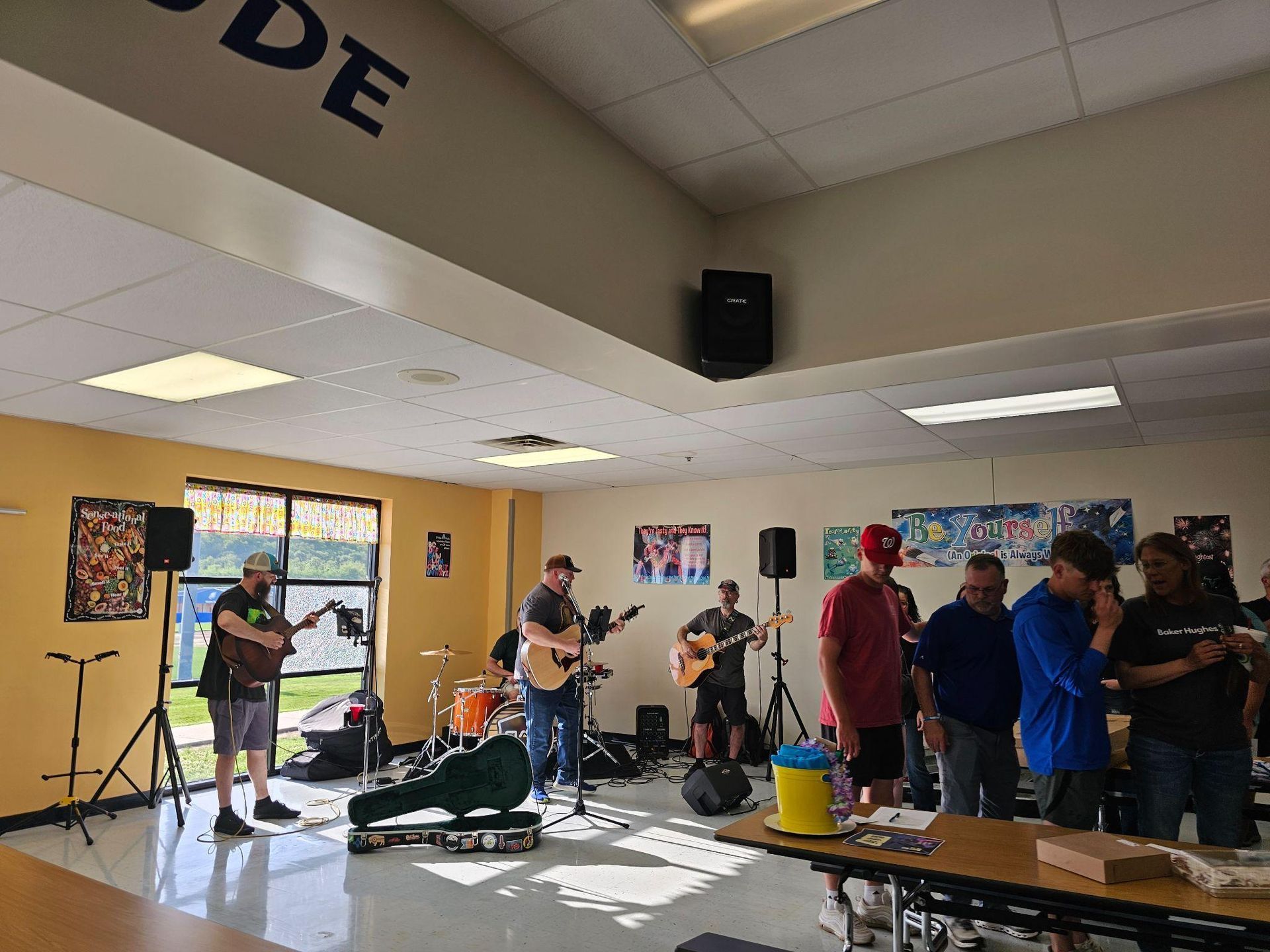 People playing instruments in a school cafeteria, with a group of onlookers standing nearby.