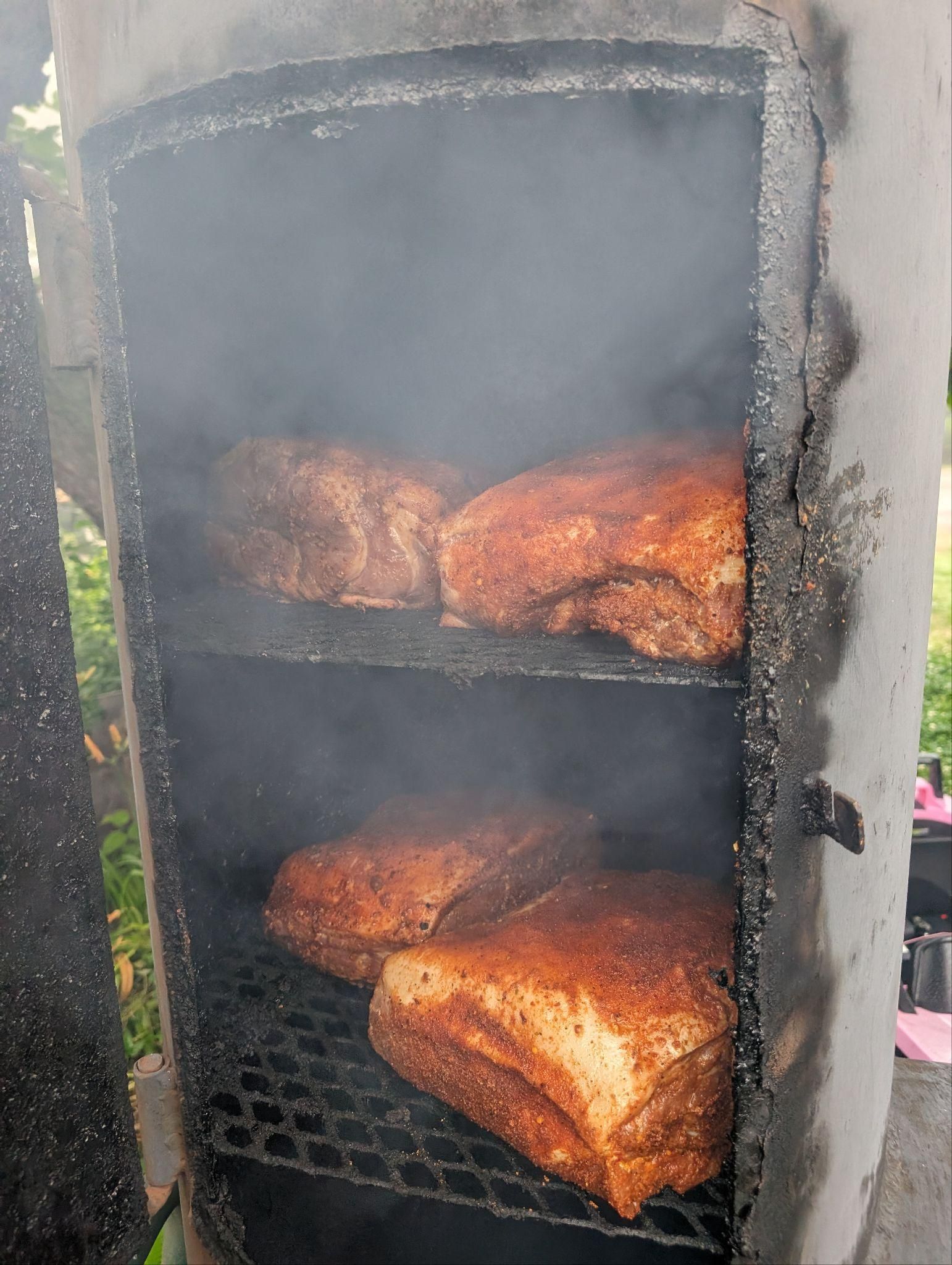 Smoked meat roasting on racks inside a vertical smoker, with smoke rising.