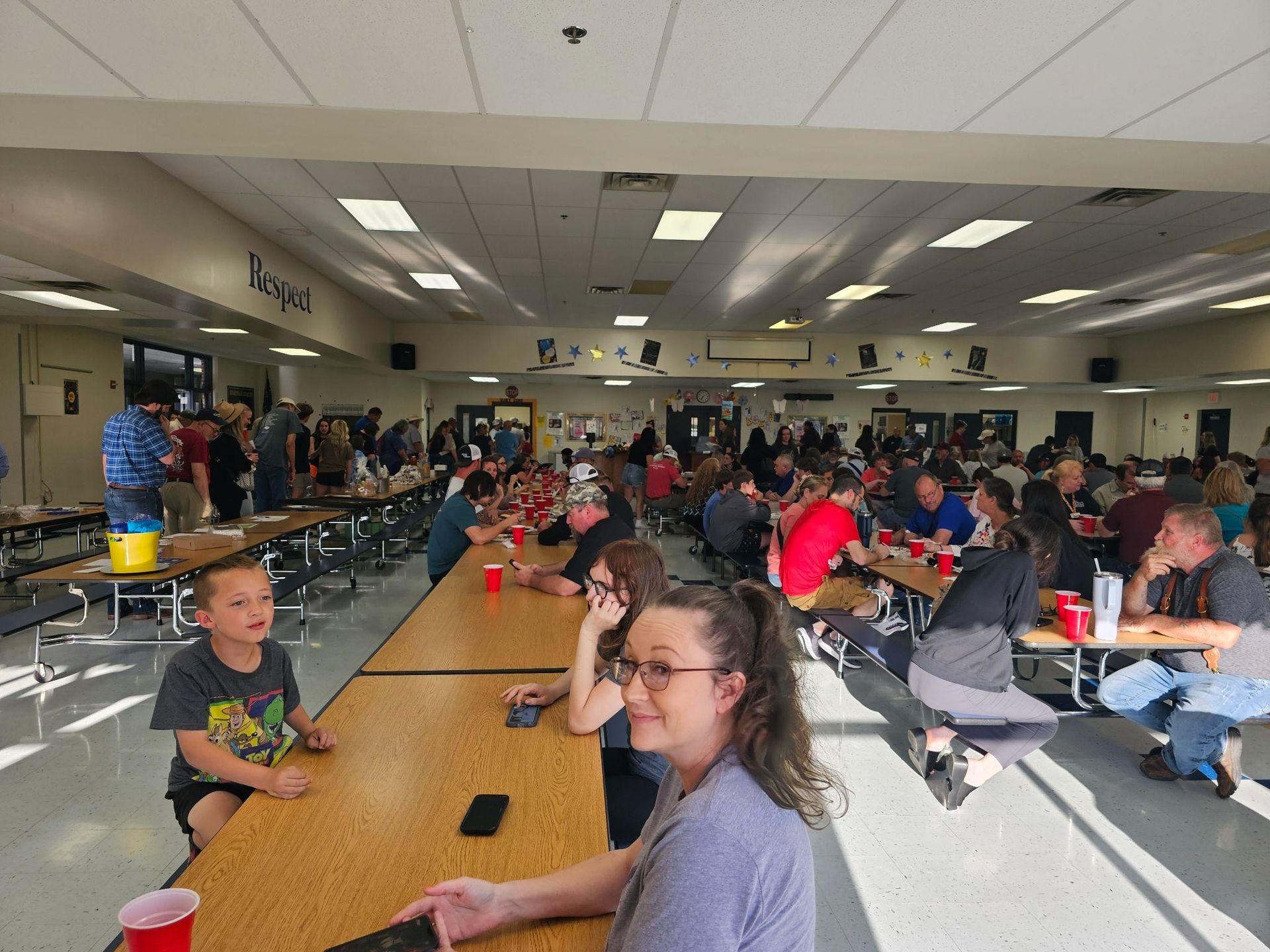 School cafeteria with many students seated at long tables, chatting and eating under bright lights.