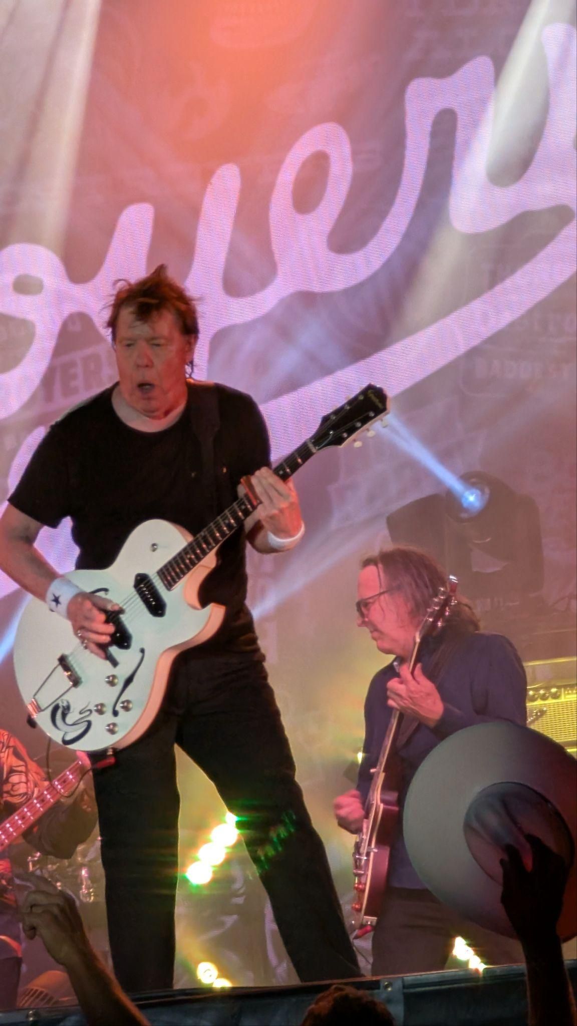 Guitarist performing onstage under pink lights, with a large white script backdrop behind him.