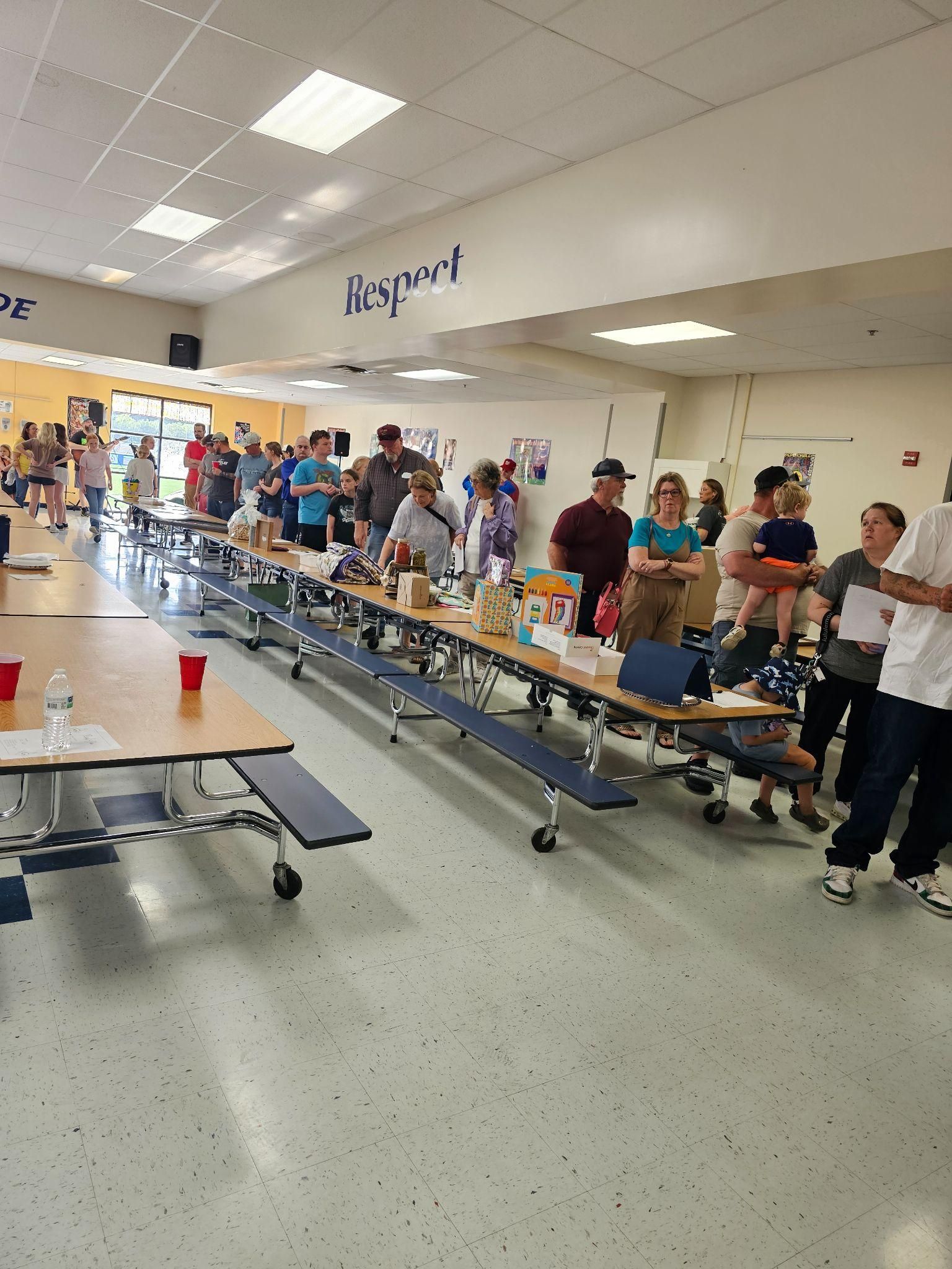 People standing along cafeteria serving tables in a bright school cafeteria, with trays and drinks on the tables.