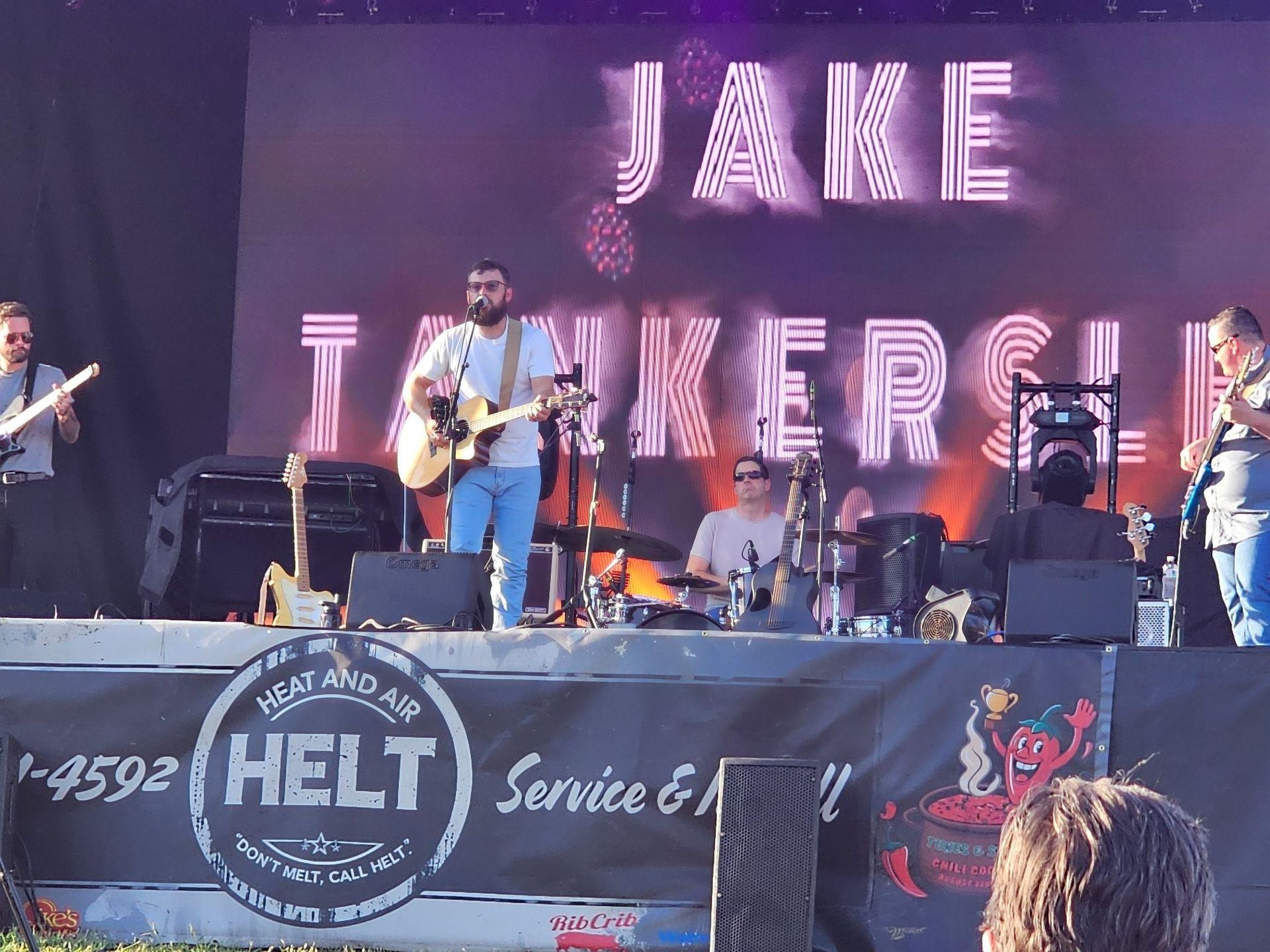 Band performing on an outdoor stage under a “JAKIE” sign, with guitars and drums.