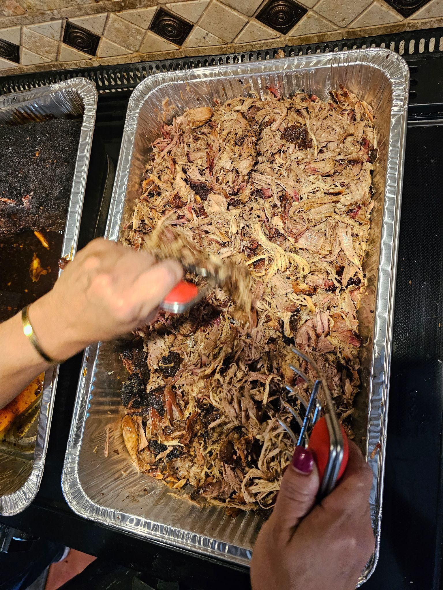 Hands serving shredded barbecue meat from a foil tray with red-tipped tongs