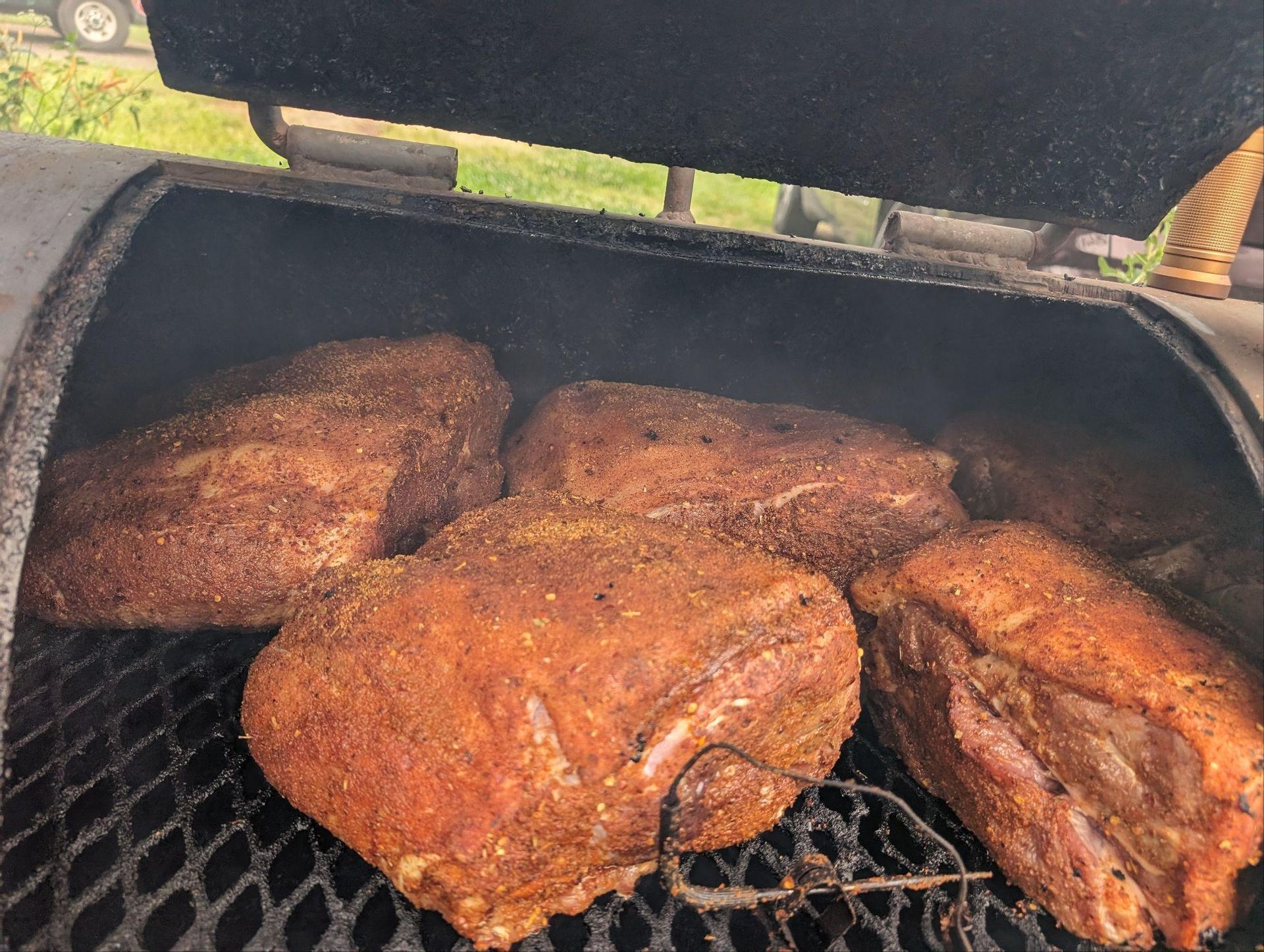 Smoked meat cuts on a grill, covered in dark seasoning and smoking outdoors