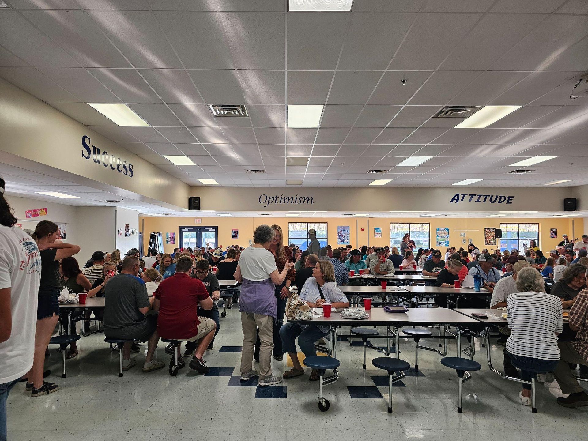 Busy school cafeteria with many people seated at long tables under bright ceiling lights