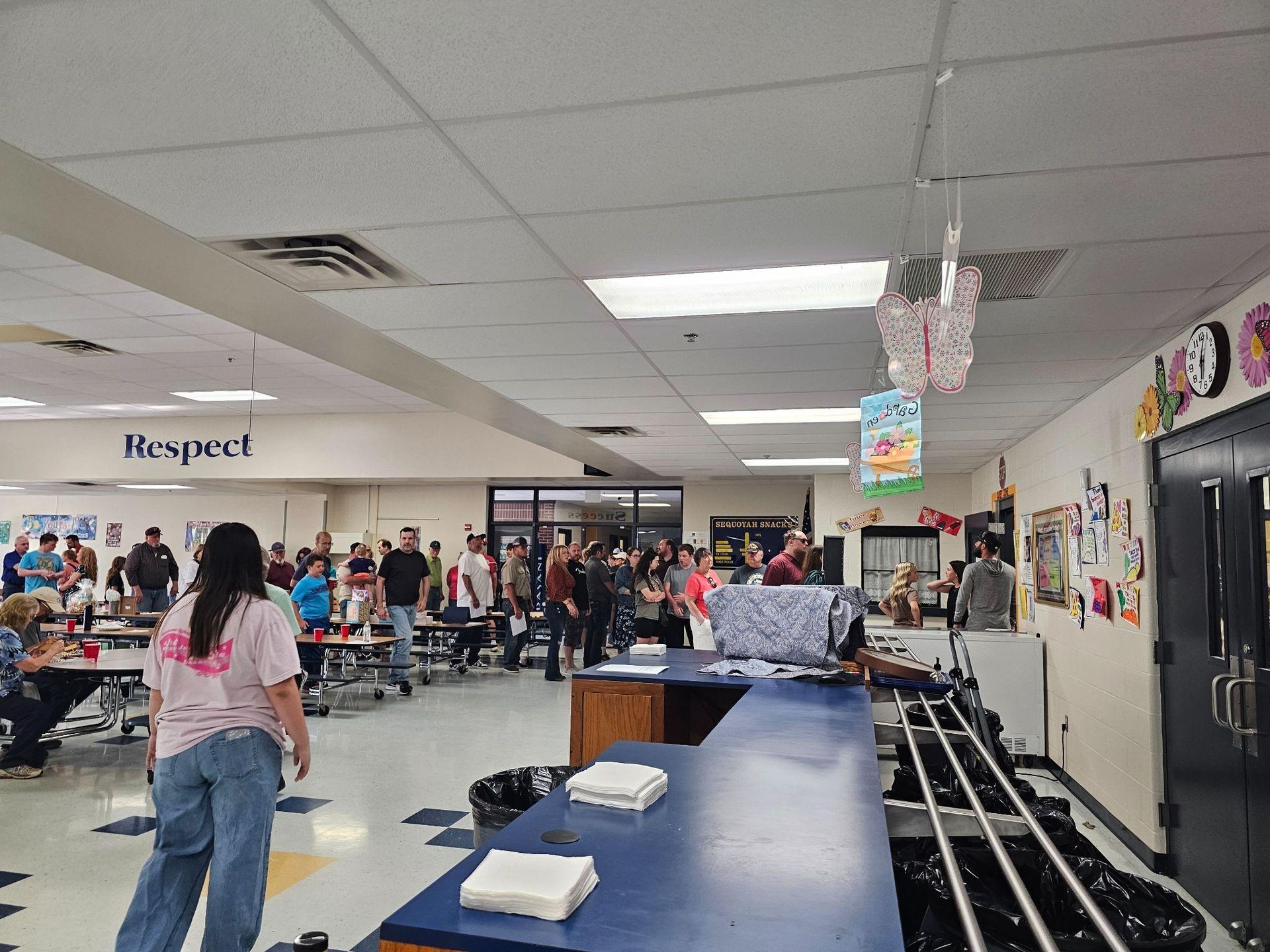School cafeteria with students standing near a serving line and tables, blue counter in foreground