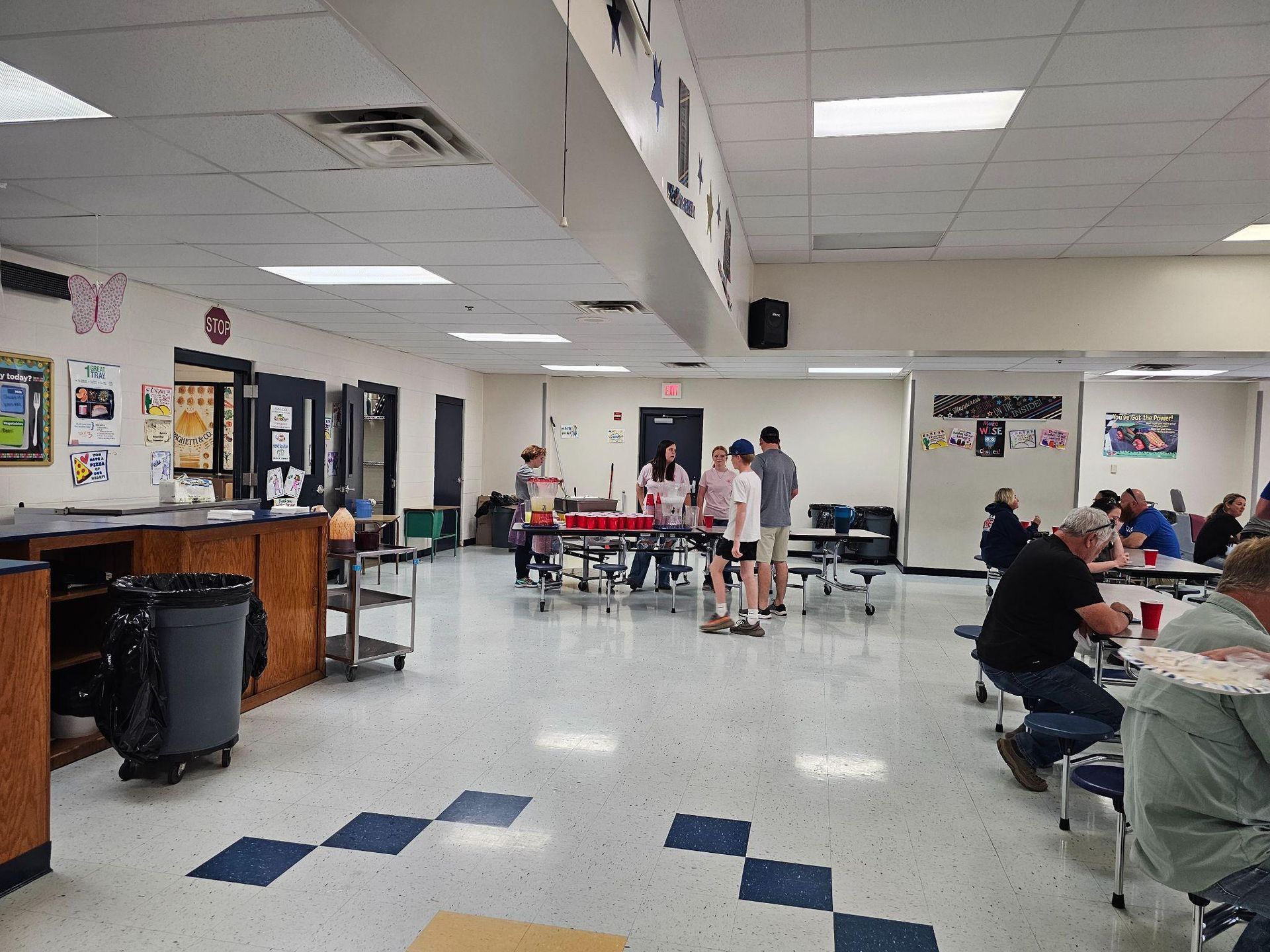 Community hall with people seated at tables and a group standing in the center under bright fluorescent lights.