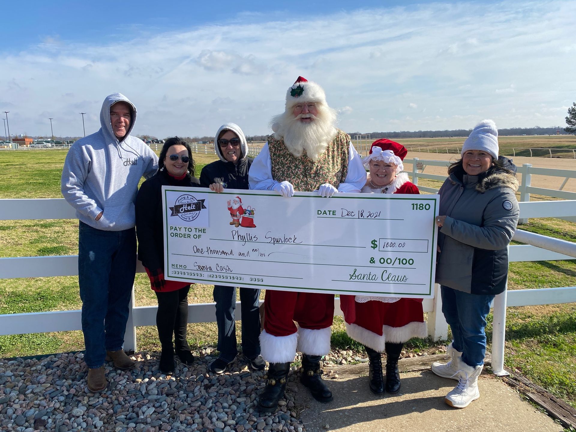 Five people outdoors holding a large check beside a person in a Santa suit on a sunny day