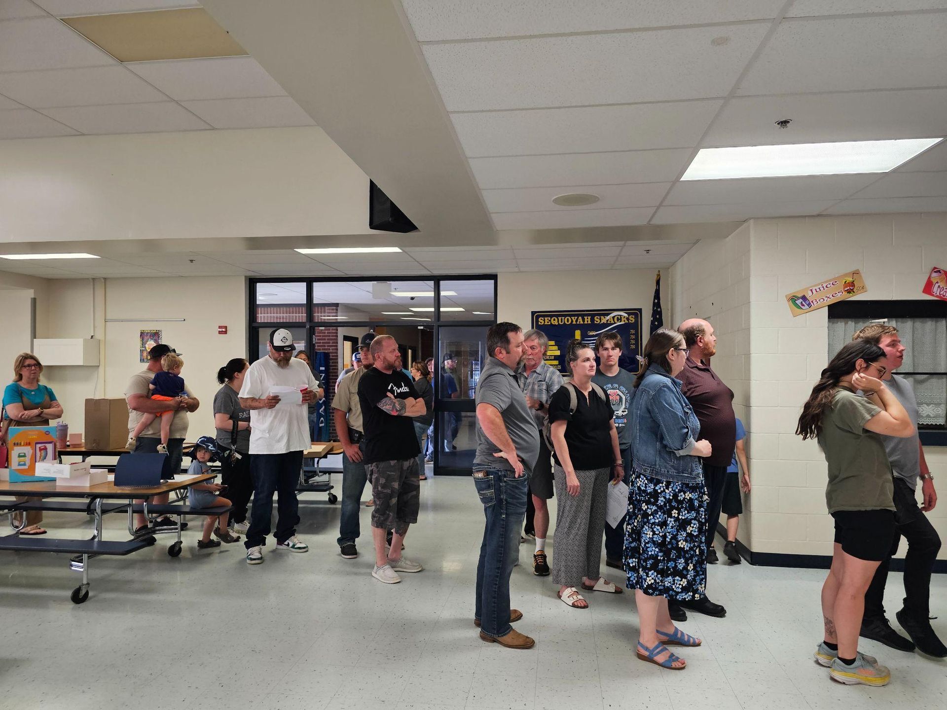 People standing in a cafeteria or community room, gathered around tables under fluorescent lights.