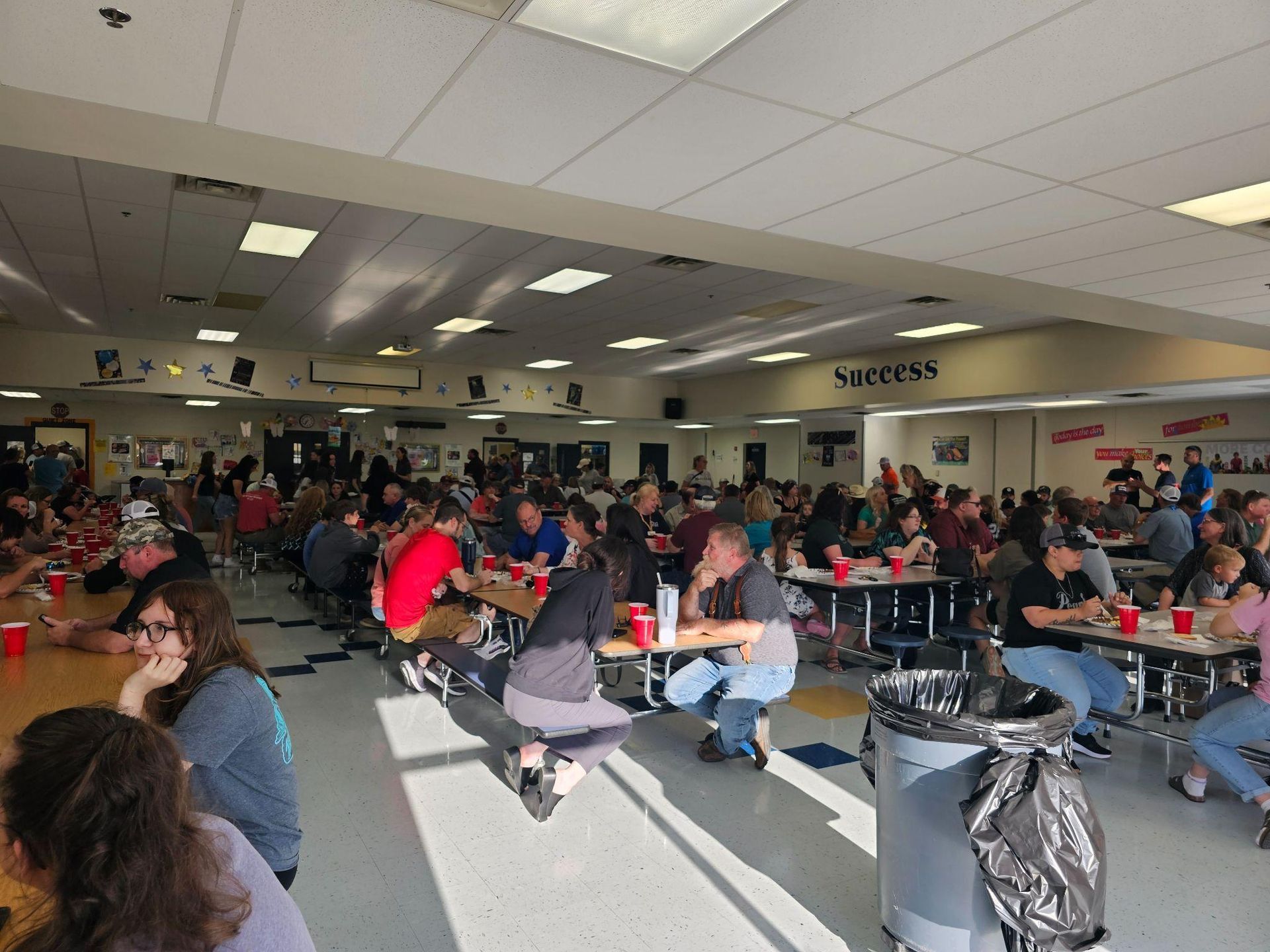 Crowded school cafeteria with students seated at long tables under bright ceiling lights