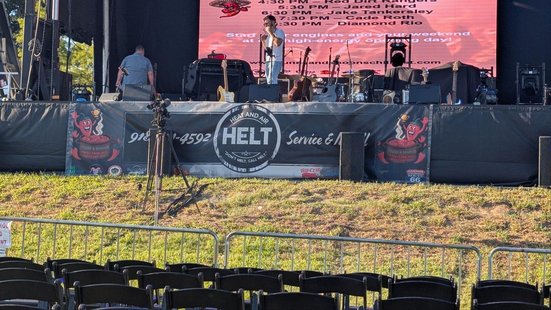Outdoor stage with musicians and a large red backdrop, viewed from a grassy field behind a black barrier.