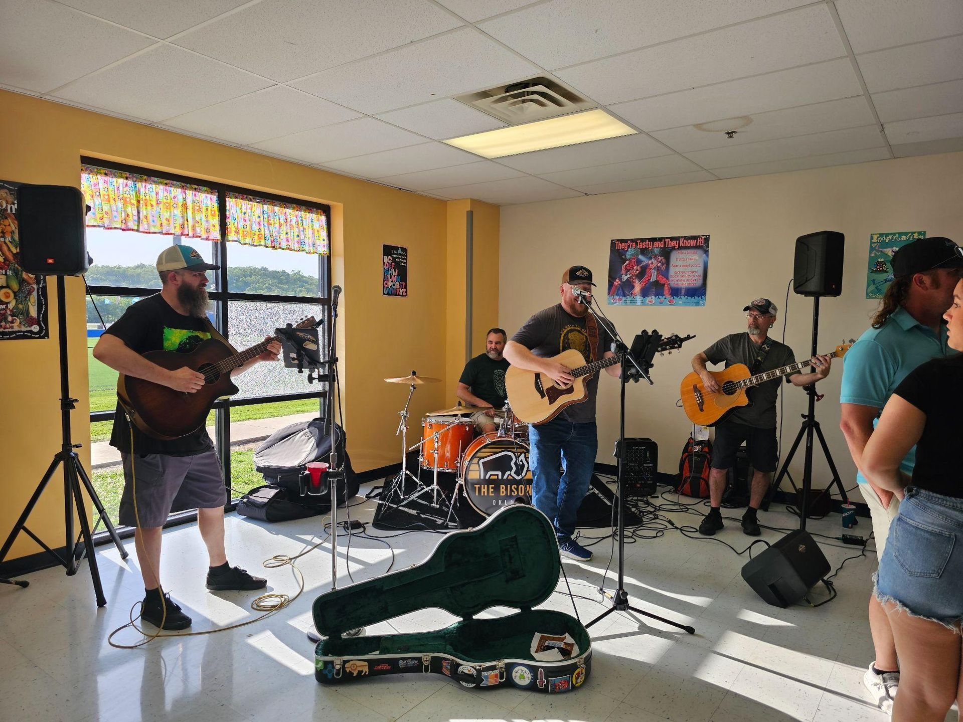 Live band rehearsing in a bright room, with guitars, drums, and a guitar case on the floor.