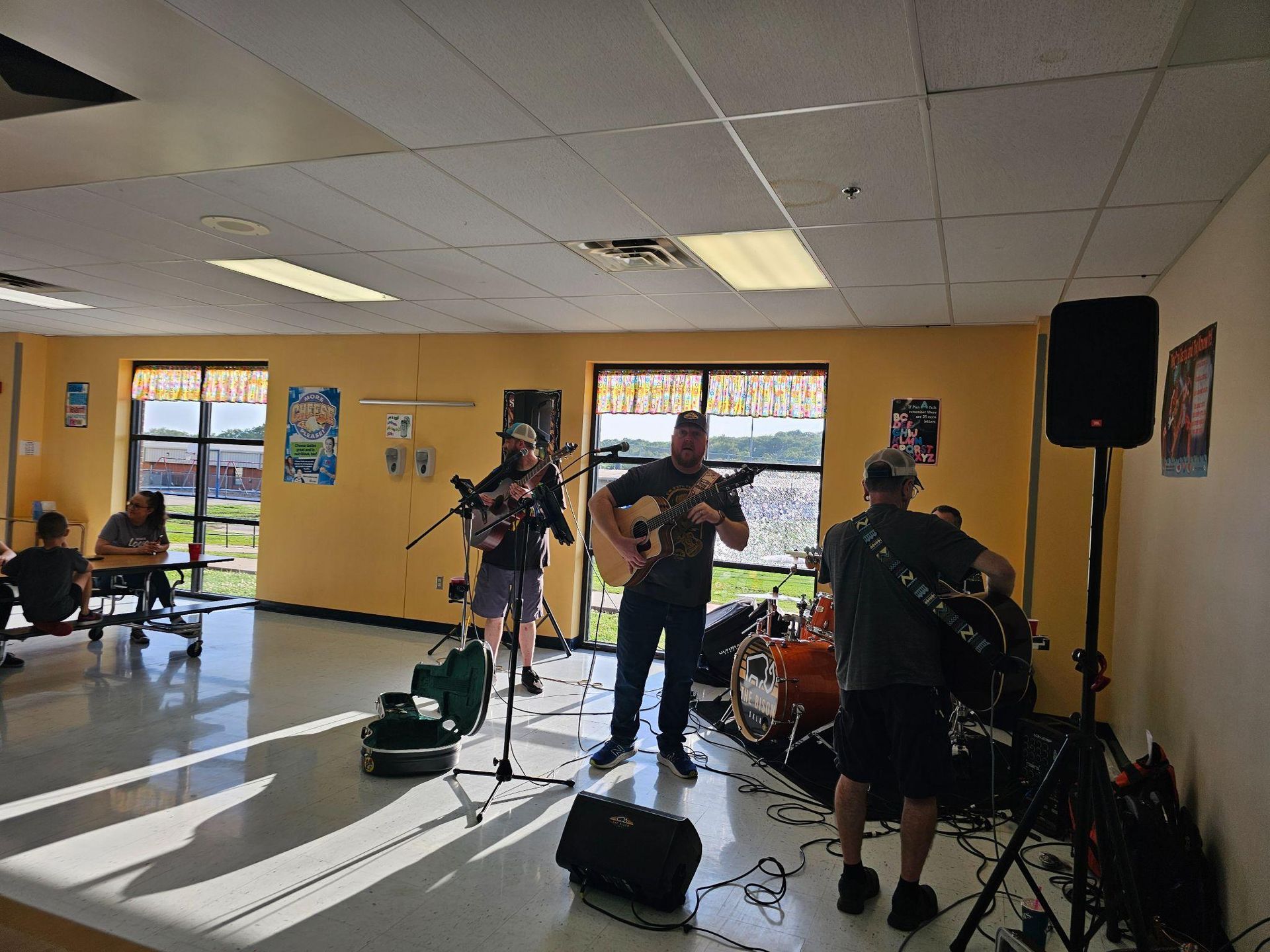 Musicians performing in a bright hall with guitars, drums, and a microphone stand.