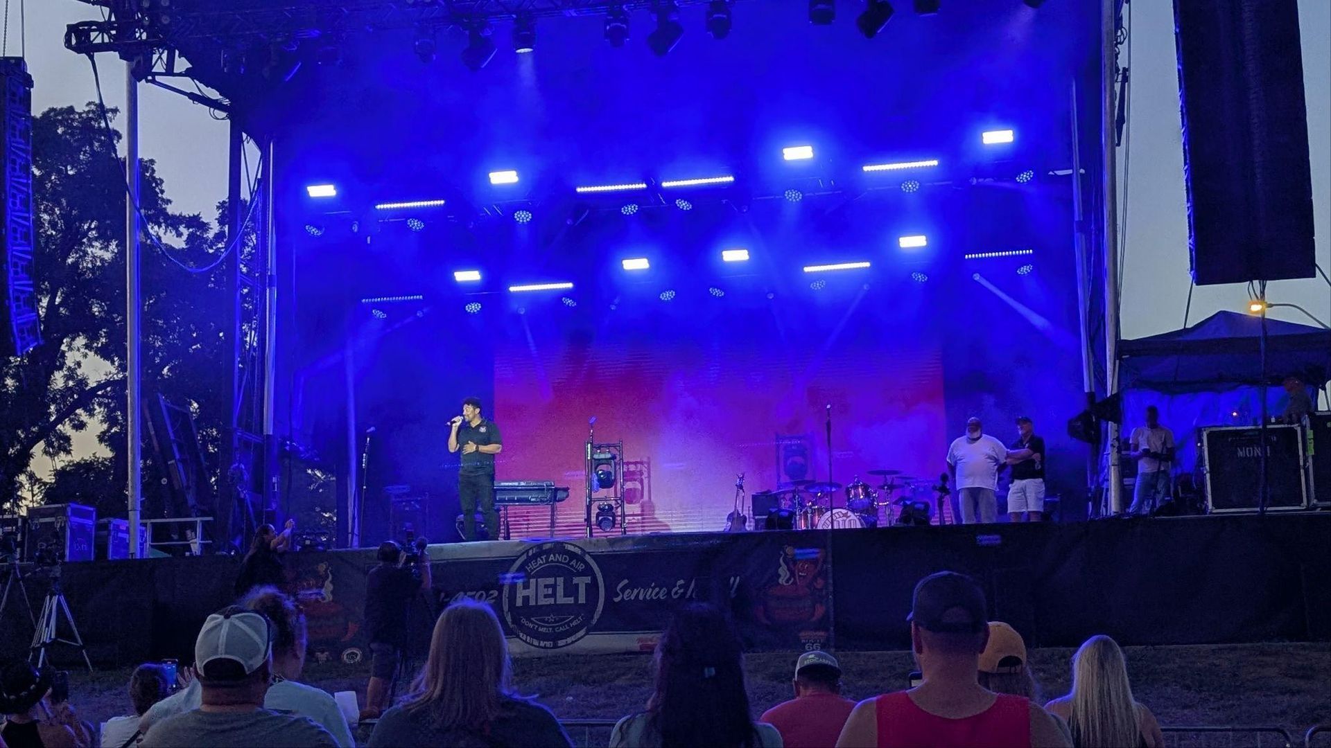 Concert stage lit blue and purple with performers and a crowd in front at an outdoor venue