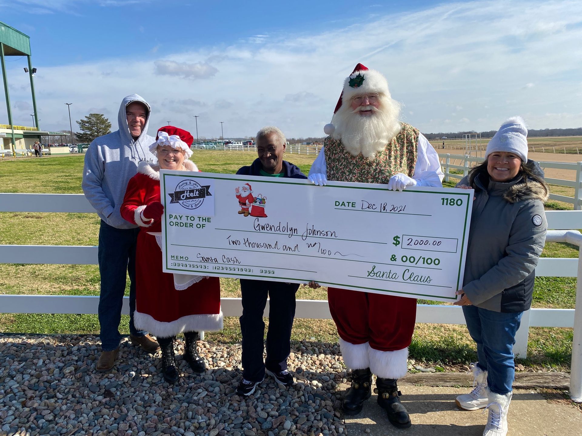 Four people in winter clothes hold a giant check outdoors on a sunny day.