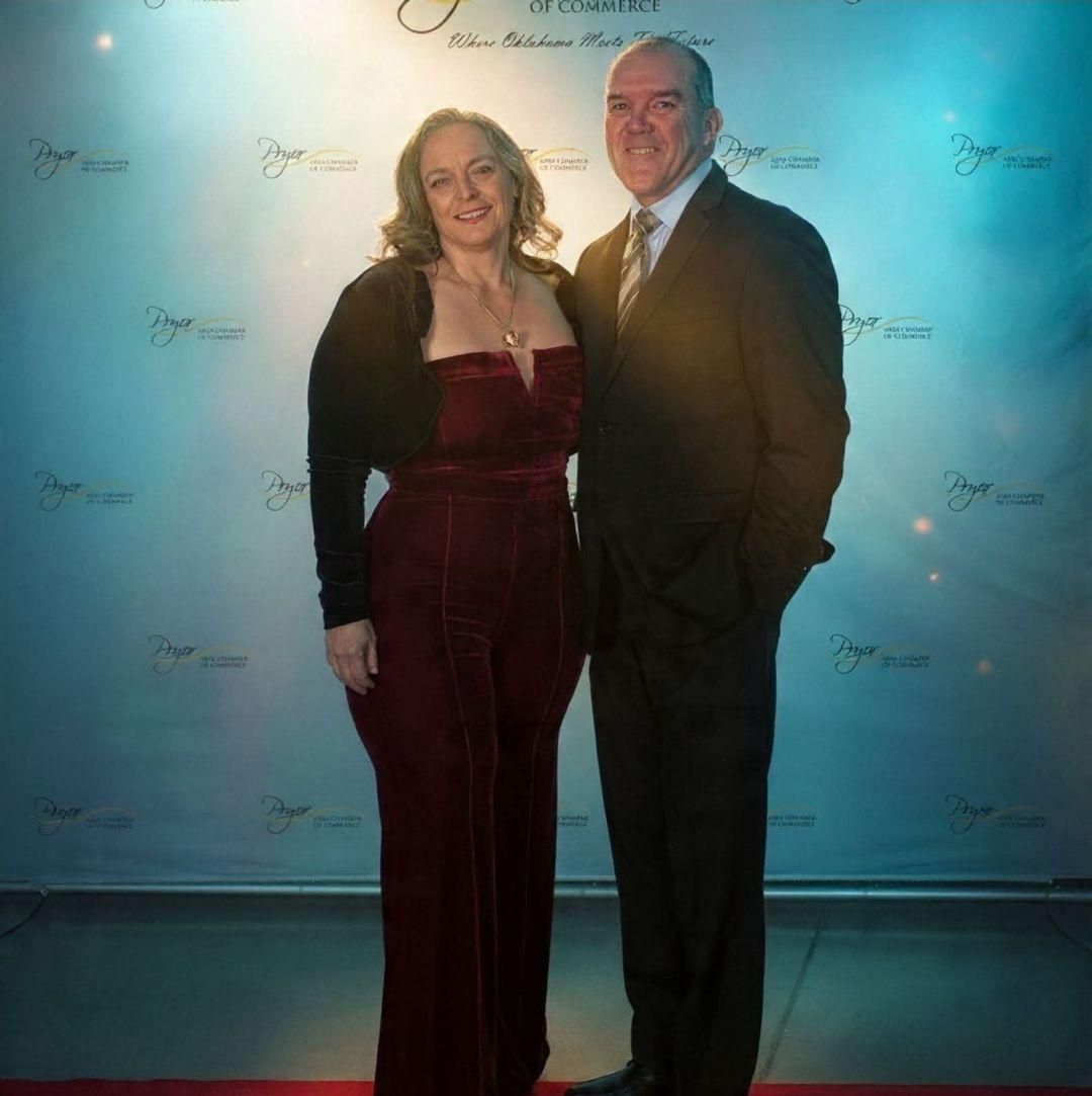 Two people posing on a blue-lit red carpet, one in a burgundy dress and one in a dark suit.